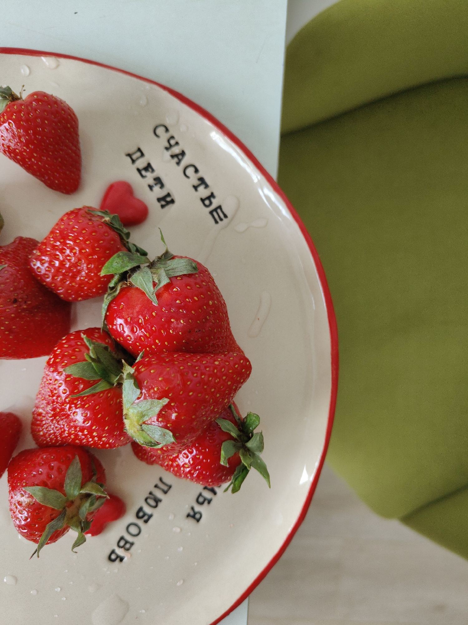 A plate of strawberries sits on a surface next to a green armchair. The plate is off-white with a red rim and Russian writing on the inside. There are several ripe, red strawberries on the plate, and a few red candy hearts. The background is blurred.