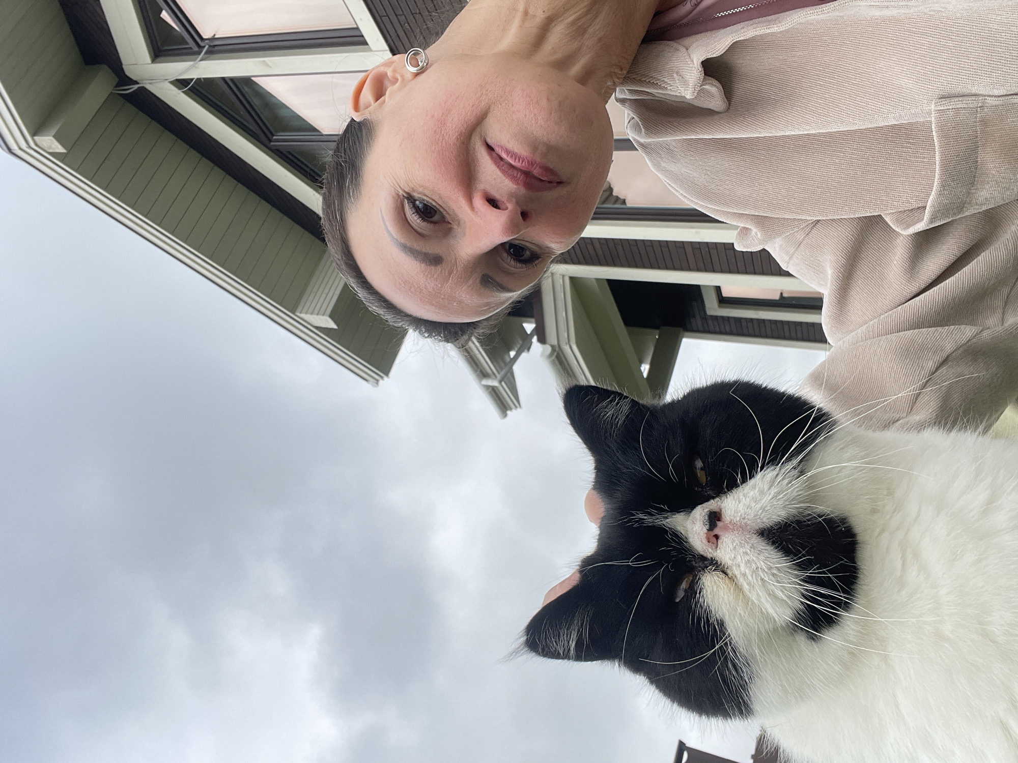 An outdoor, waist-up, eye-level, medium shot features a woman and a cat against a backdrop of a house and cloudy sky. The woman is in the right foreground, with her head tilted down towards the cat and her face turned to the right, looking at the camera with a slight smile and her eyes open. She has light skin, dark hair pulled back, and is wearing a light-colored corduroy jacket over a pink top. She has a silver circular earring in her right ear. A black and white cat is in the left foreground, with its face and chest visible. Its eyes are narrowed, and it appears to be looking down. Its fur is black and white, with black markings on its face. The cat's right ear is being gently touched by the woman's hand. The house behind them has a dark brown facade with white trim, and green-painted eaves. The sky is overcast and grey. The location is identified as the city of Solov'evka, Russia.