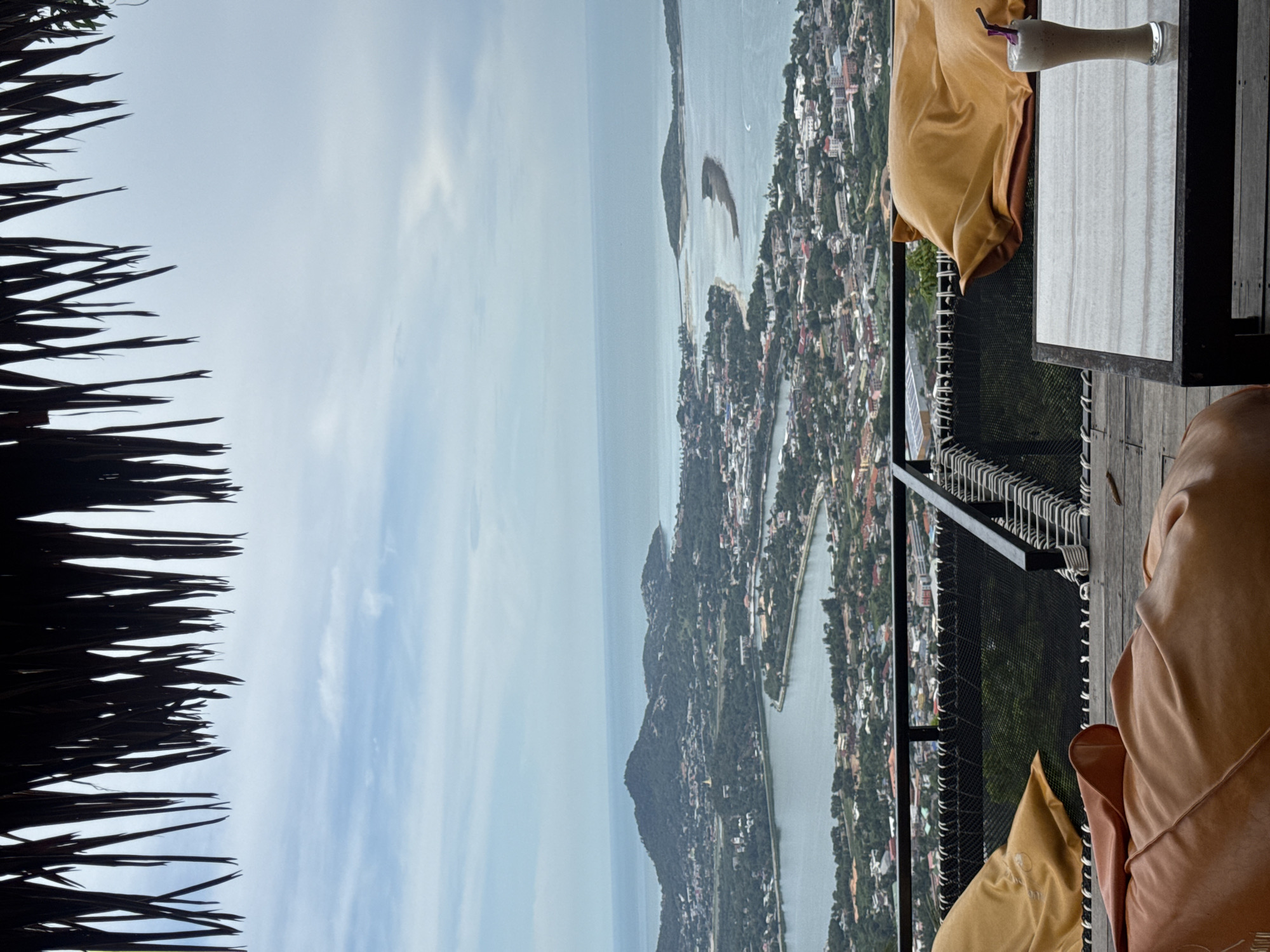 A wide-angle, elevated shot shows a serene view of a coastal town and the ocean, with a thatched roof overhang in the foreground. Two comfortable, oversized beanbag chairs, one in a muted orange and the other in a golden yellow, are placed on a wooden deck, offering a prime spot for relaxation. A glass of iced coffee with a straw sits on a low, dark-colored table, adding a touch of leisure to the scene. The town sprawls along the coastline, with buildings nestled among lush greenery, and beyond it, the calm blue ocean stretches to the horizon under a cloudy sky. The scene evokes a sense of tranquility and a vacation atmosphere.