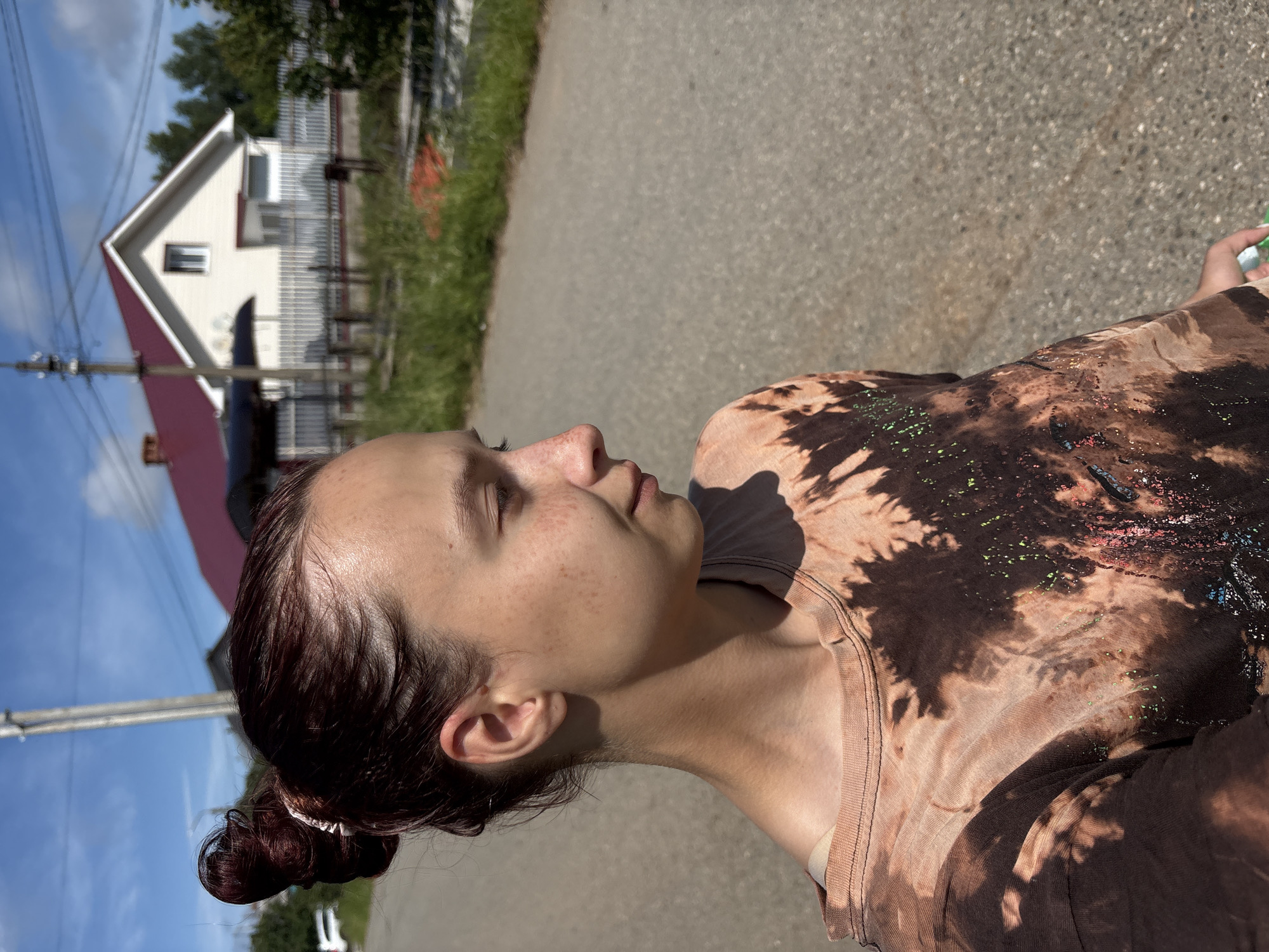 A young woman with dark hair pulled into a bun stands on an asphalt road on a sunny day. She is facing to the right of the frame and looking away from the camera. Her face is lightly tanned and she has freckles across her nose and cheeks. She is wearing a short-sleeved t-shirt with a tie-dye pattern in shades of brown and tan, with some green and red splatters on the front. In the background, there is a white house with a red roof, a white picket fence, green grass, and trees. A utility pole with wires is visible to the left of the house. The sky is blue with some white clouds. The location is likely a rural or suburban area in the city of Прикамский, Russia.