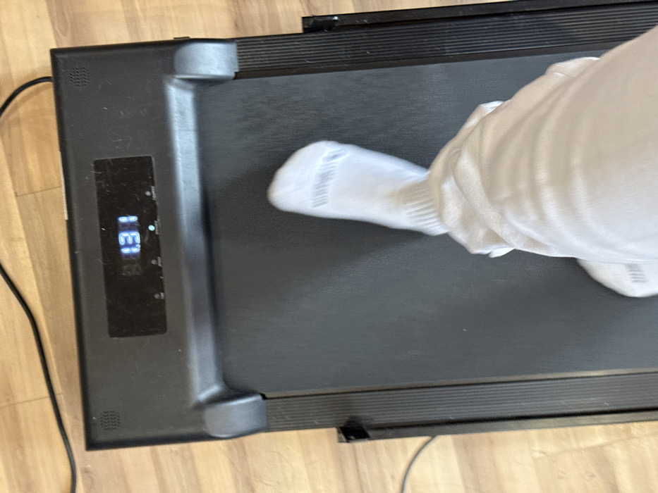 The scene depicts an overhead view of a person operating a black treadmill indoors, located in Galabovtsi, Bulgaria. The treadmill's digital display shows the number "131" and the letters "HI" illuminated in blue. A segment of a power cord is visible on the light-colored wooden floor extending from the top-left of the image. The lower part of a person's right leg and foot, clad in a white athletic sock and light-colored athletic pants, is visible centered on the treadmill's black running surface, indicating motion. The treadmill features textured black side rails.