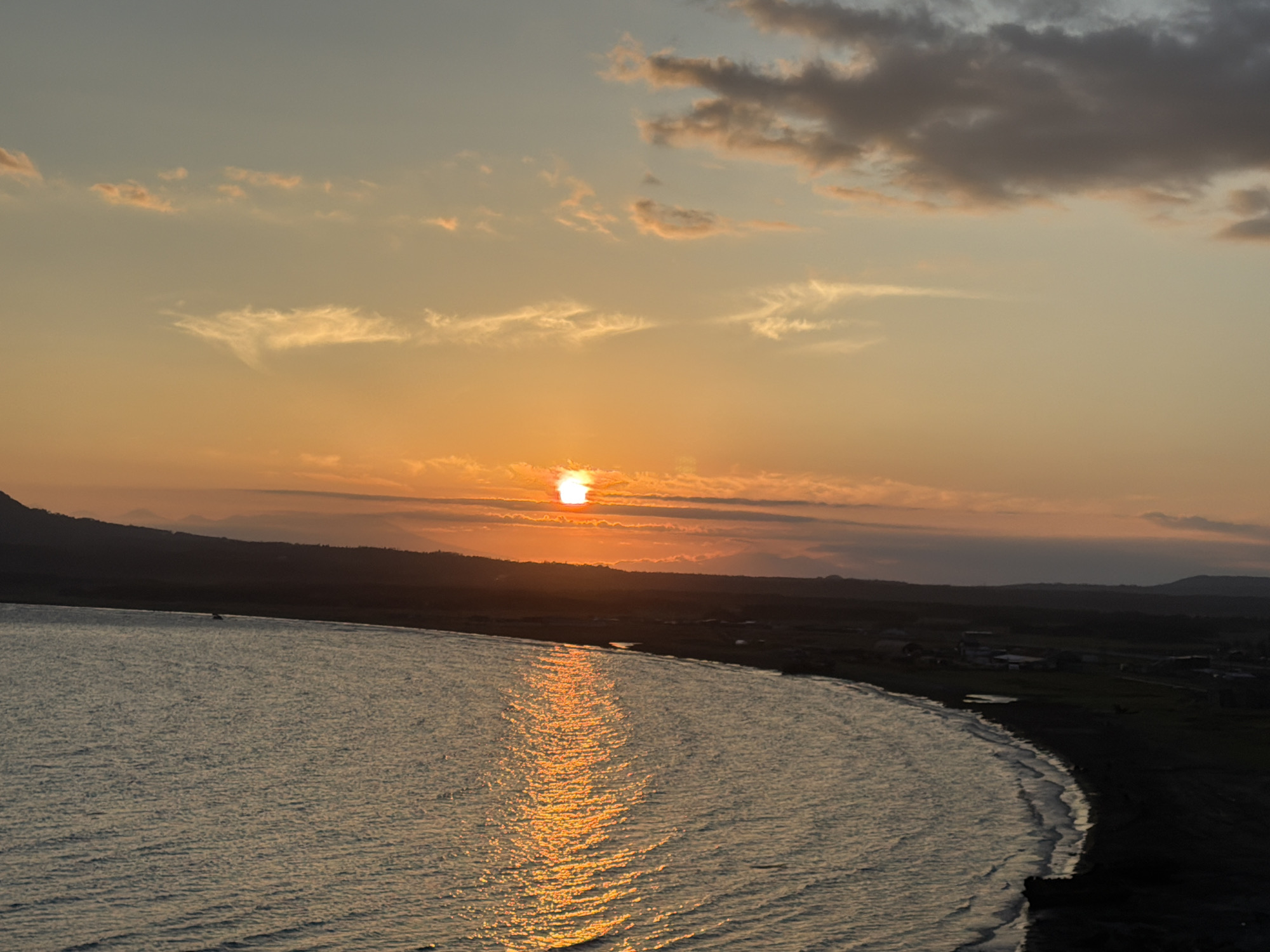 The image depicts a sunset over a body of water near Yuzhno-Kurilsk, Russia. The sun, a bright orange orb, is partially obscured by thin clouds and is setting behind a distant, dark-hued mountain range. The sky exhibits a gradient of colors, transitioning from a pale blue at the top to warm oranges and yellows near the horizon. Wispy clouds are scattered across the sky, catching the sunset's light. The water in the foreground is calm, with ripples reflecting the golden light of the setting sun, creating a shimmering path. Along the far shore, a dark, undulating landmass is visible, sparsely dotted with what appear to be small buildings or structures, suggesting a coastal settlement. The overall scene is serene and highlights the natural beauty of the coastline during twilight.