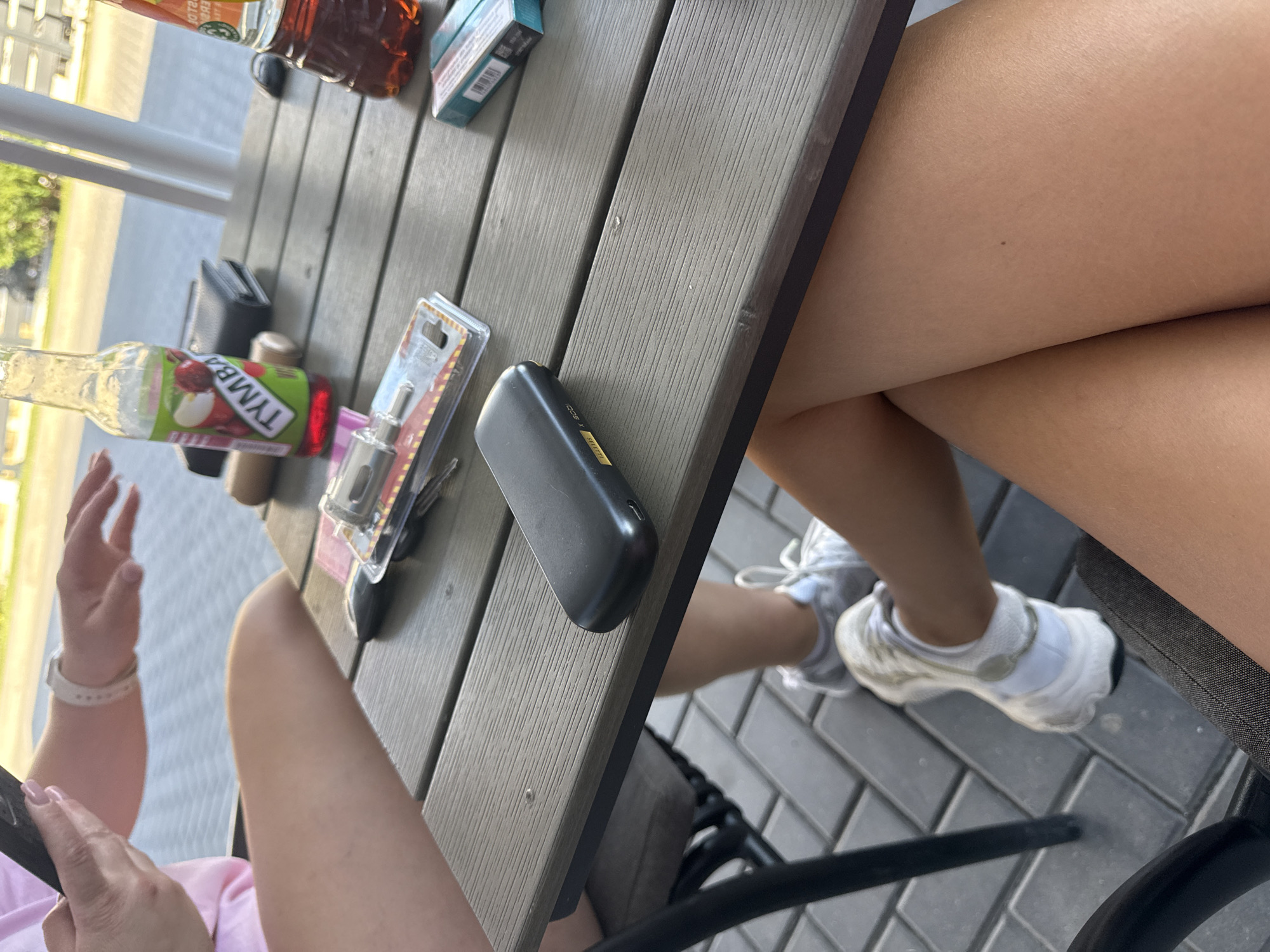 The image shows a close-up view of a person's legs and a table at an outdoor cafe, likely in Comrat, Moldova. On the table are various items including a smartphone, a black wallet, a bottle of "TYMBA" brand drink, a pack of cigarettes, and a black electronic device. The background suggests a paved outdoor seating area with chairs. The lighting indicates it is daytime, possibly late afternoon or early evening.