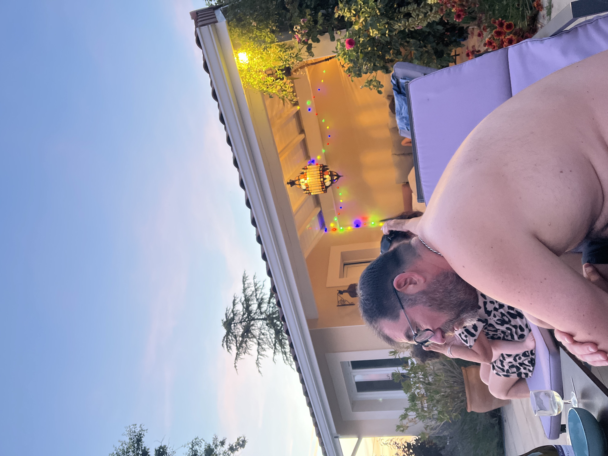 A group of people are gathered outdoors in the city of Saint-Galmier, France, likely at a residence, as indicated by the house and patio furniture. In the foreground, a man with glasses and a beard is seen from the shoulders up, facing to the left. He is shirtless and appears to be resting on a purple lounge chair. To his left, a glass with white wine and a blue bowl are on a dark table. Behind him, a woman in a leopard-print dress is seated on a lounge chair, and another person is visible behind her. The background features a house with a covered patio, decorated with string lights and a Moroccan-style lantern. Lush greenery and flowering plants surround the area, suggesting a warm climate and likely evening or dusk hours given the ambient lighting and sky.