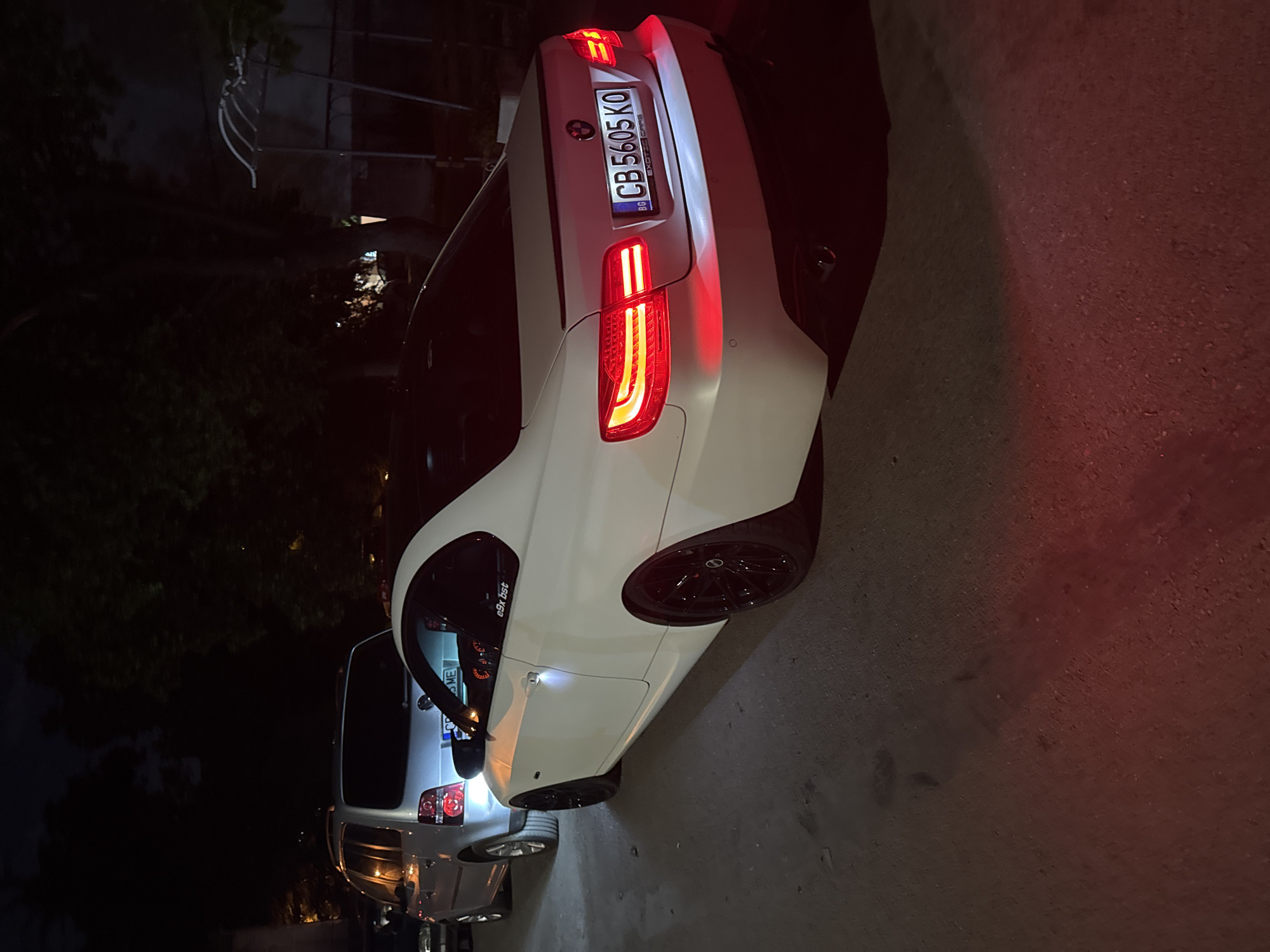 A nighttime scene shows a white BMW coupe with Bulgarian license plate "CB 5605 KO" parked on an urban street. The car's taillights are illuminated. Another vehicle, possibly a silver SUV, is partially visible behind the BMW. The environment is darkly lit, with street lighting visible in the background. The scene is located in Sofia, Bulgaria based on the license plate origin.