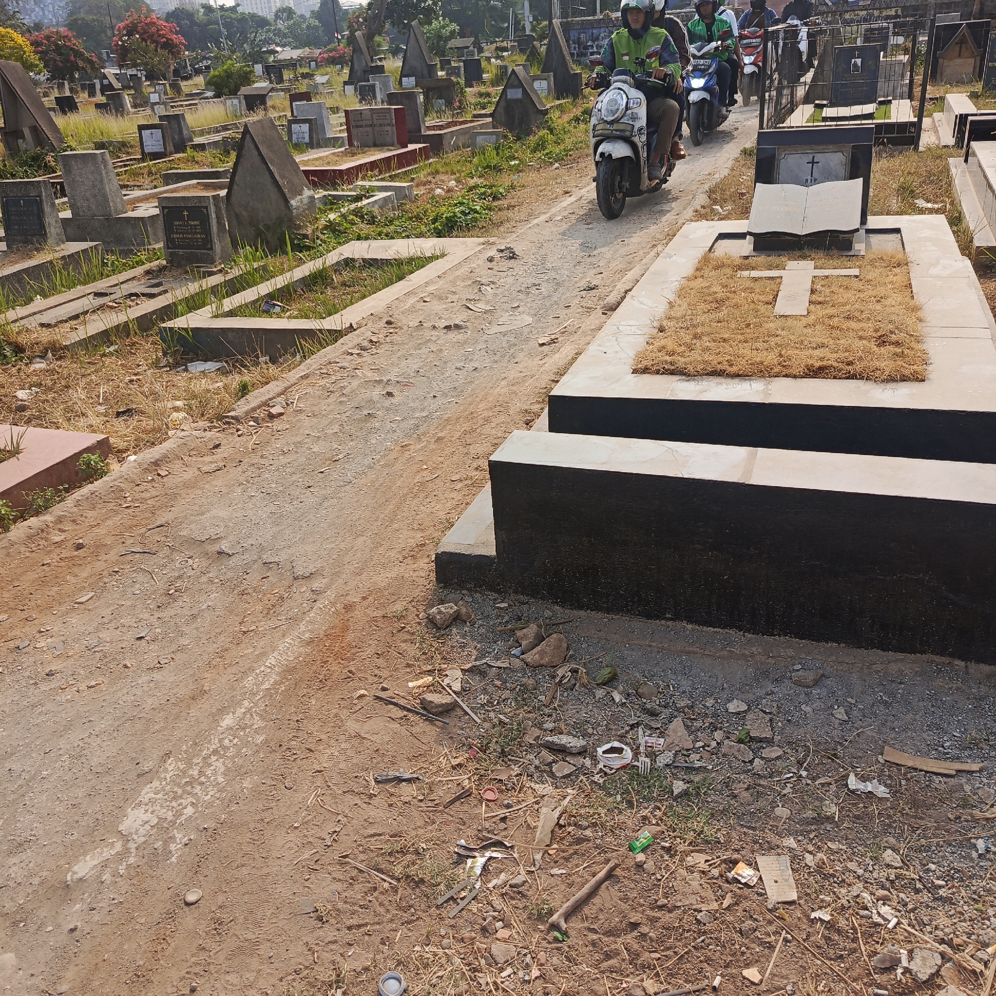 In South Jakarta, Indonesia, this image captures a cemetery with various gravestones and monuments, some adorned with crosses. A dirt path winds through the grounds, with evidence of tire tracks suggesting recent vehicle passage. On the path, several individuals are riding motorcycles, navigating the uneven terrain. The surrounding environment includes overgrown grass and scattered debris, with some gravestones exhibiting signs of age and neglect. The presence of buildings in the background indicates the cemetery is situated within an urban area.