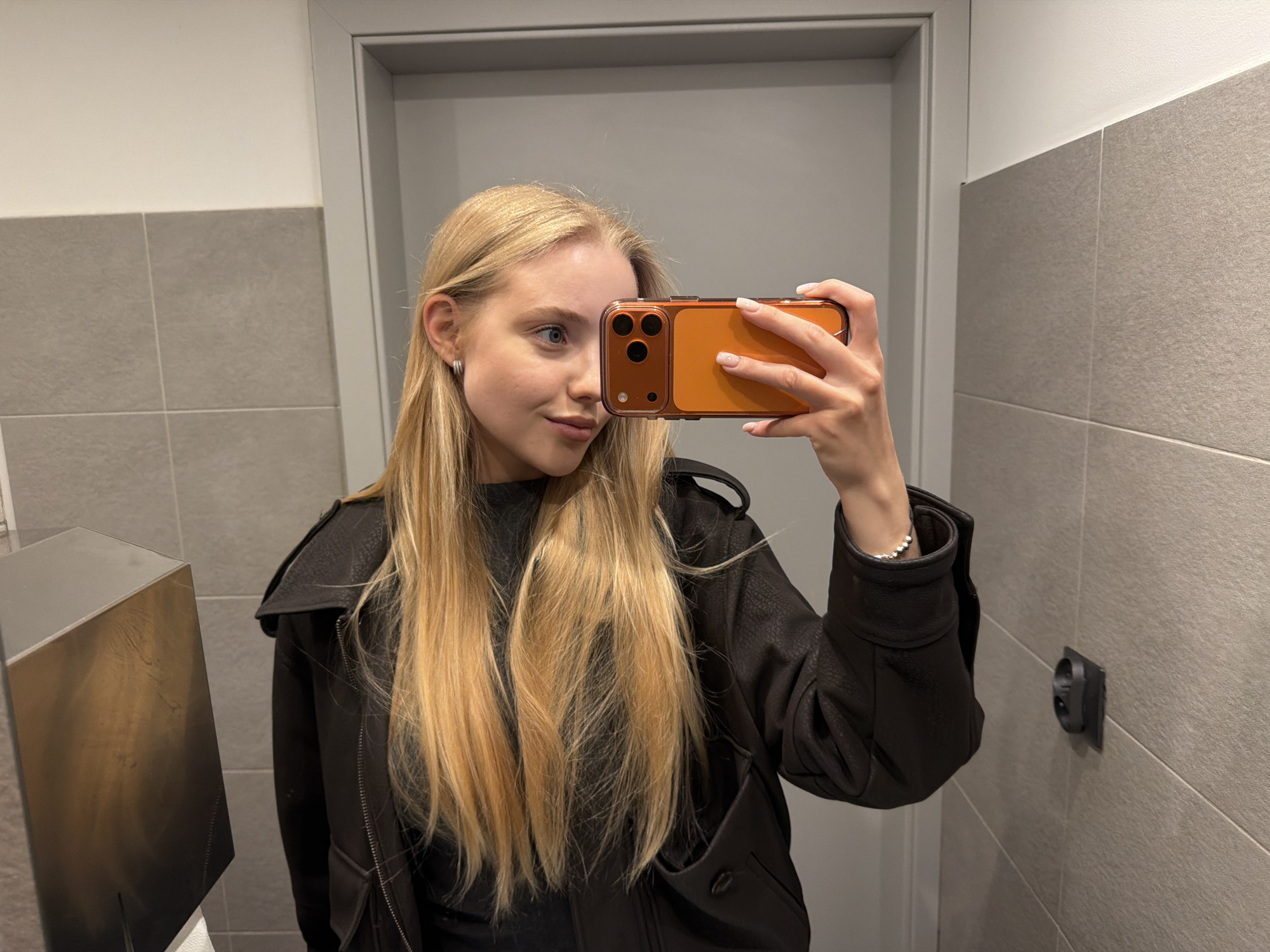 A young woman with long blonde hair, wearing a black jacket and dark top, is taking a selfie in what appears to be a bathroom. She is holding an orange-cased phone with her right hand, obscuring part of her face. She is standing in front of a closed, grey door. The wall to her left is covered with grey tiles, and a metal dispenser is visible. To her right, the tiled wall features an electrical outlet. The location is Unknown, Unknown.