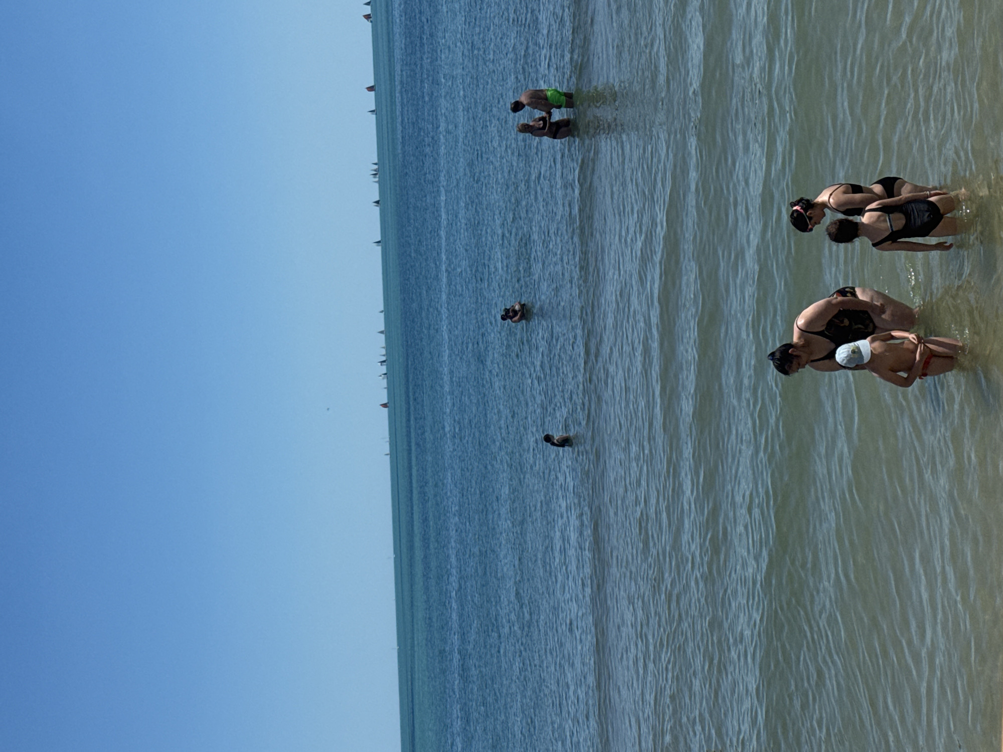 The image depicts a group of people enjoying a day at the beach in Varna, Bulgaria. The scene is set on a sandy shore, with the calm, clear waters of the Black Sea stretching out to the horizon. The sky is a vibrant, cloudless blue, suggesting a sunny and pleasant day, likely late morning or early afternoon.
Several individuals are visible in the water, engaged in various activities. In the foreground, a family or group of friends appears to be interacting. A woman in a black bikini is leaning over, seemingly assisting a child wearing a white cap and red bottoms. Beside them, another woman in a black swimsuit is also bent over, possibly playing with the child or observing them. Further out in the water, a couple stands together, with one person wearing green shorts and the other in a dark swimsuit. Another individual is seen further back, partially submerged in the water.
The water is shallow near the shore, gradually deepening as it recedes. The sunlight creates shimmering patterns on the surface of the water, highlighting the gentle ripples. In the distance, a line of small boats or buoys can be seen dotting the sea, adding to the sense of a popular recreational area. The overall atmosphere is one of leisure and enjoyment, with people taking advantage of the warm weather and coastal setting. No visible text is present in the image.