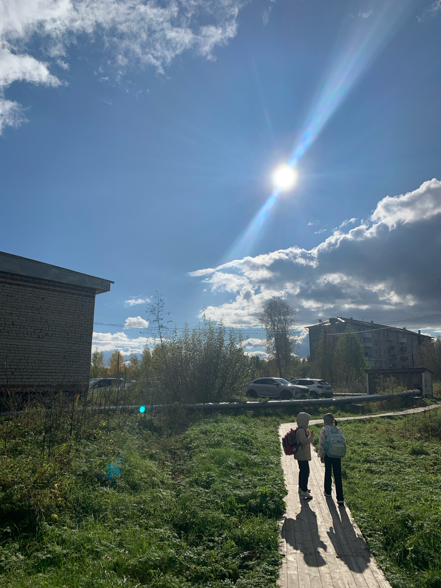 The image depicts a sunlit outdoor scene in Vas'kovo, Russia. Two young children, one wearing a light brown hooded jacket and a red backpack, and the other wearing a patterned jacket and a light blue backpack, are walking away from the camera on a wooden walkway. They are casting long shadows on the walkway. The environment is characterized by lush green grass on either side of the walkway, with some trees and bushes in the background. To the left, a low-rise building constructed of grey brick is visible. Further in the background, an apartment building and several parked cars are present. The sky is a bright blue with scattered white and dark grey clouds, and the sun is prominently featured with a visible lens flare.