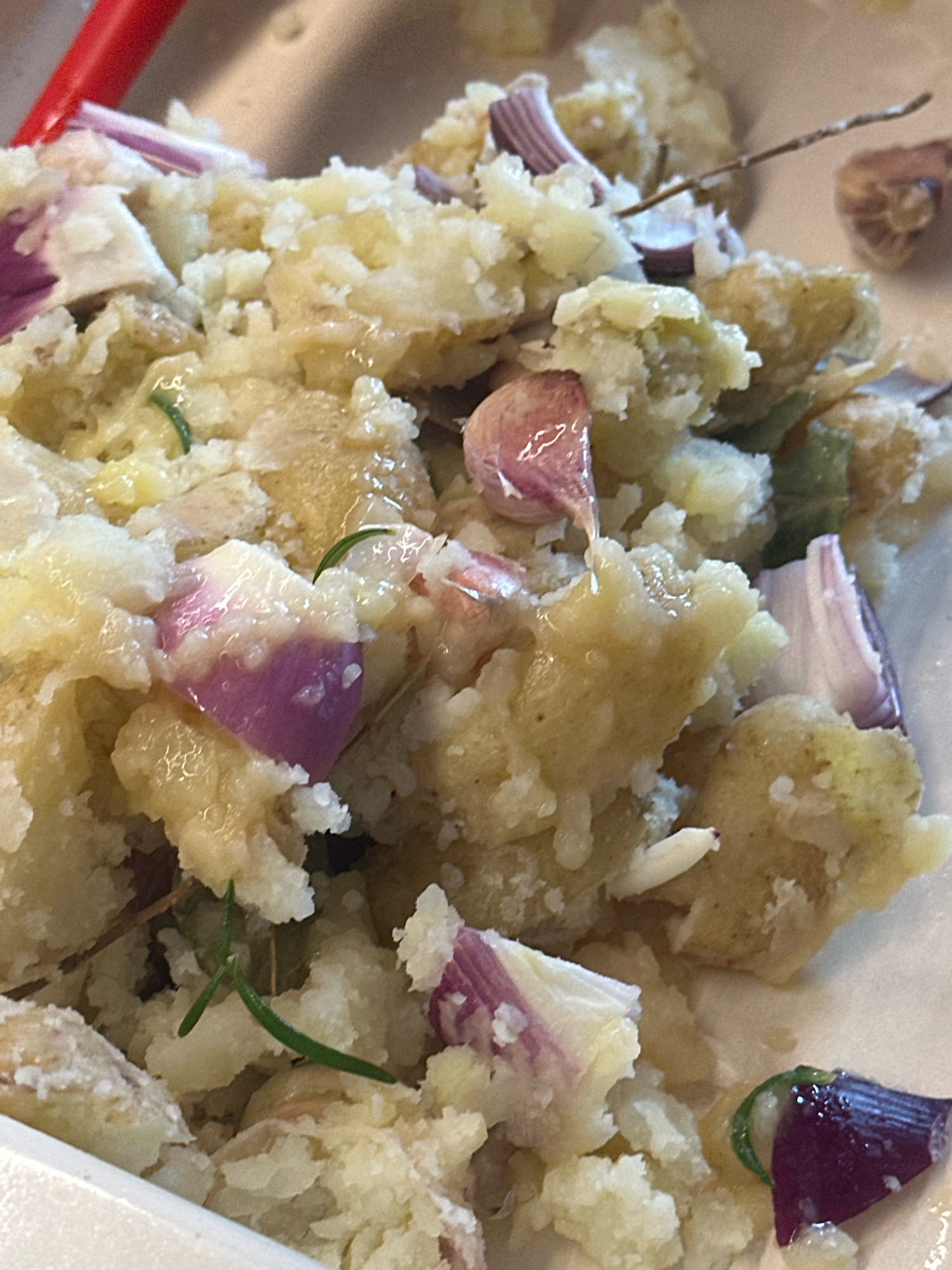 This is a close-up, overhead shot of a dish that appears to be smashed potatoes mixed with red onion and garlic. There are sprigs of rosemary throughout the dish. The lighting is bright, and the dish is served in a white container. There is a red utensil visible on the left side of the image. The scene is a kitchen or dining area. No specific location within Nevinnomyssk, Russia, can be determined from the image.