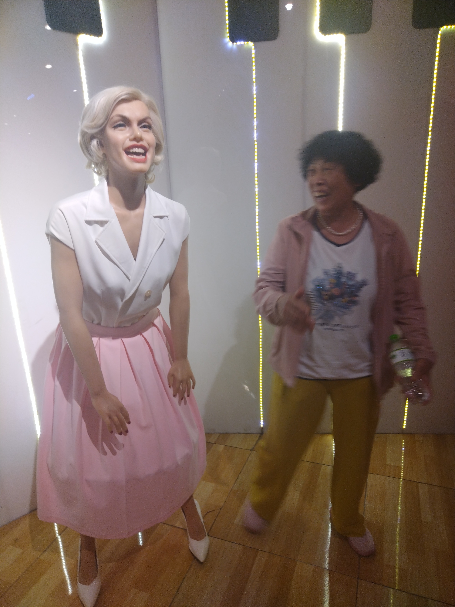 The image depicts a woman standing next to a wax figure resembling Marilyn Monroe. The woman is on the right side of the frame, wearing a pink jacket, a white T-shirt with a floral design, yellow pants, and pink shoes. She is holding a bottle of water in her right hand and gesturing with her left hand. The wax figure is on the left side, wearing a white top with a pink skirt and white shoes. The figure has a blonde hairstyle and is smiling. The background is a white wall with vertical light strips. The scene appears to be indoors, possibly in a wax museum. The location is noted as Unknown, Unknown. The floor is tiled with a shiny finish.