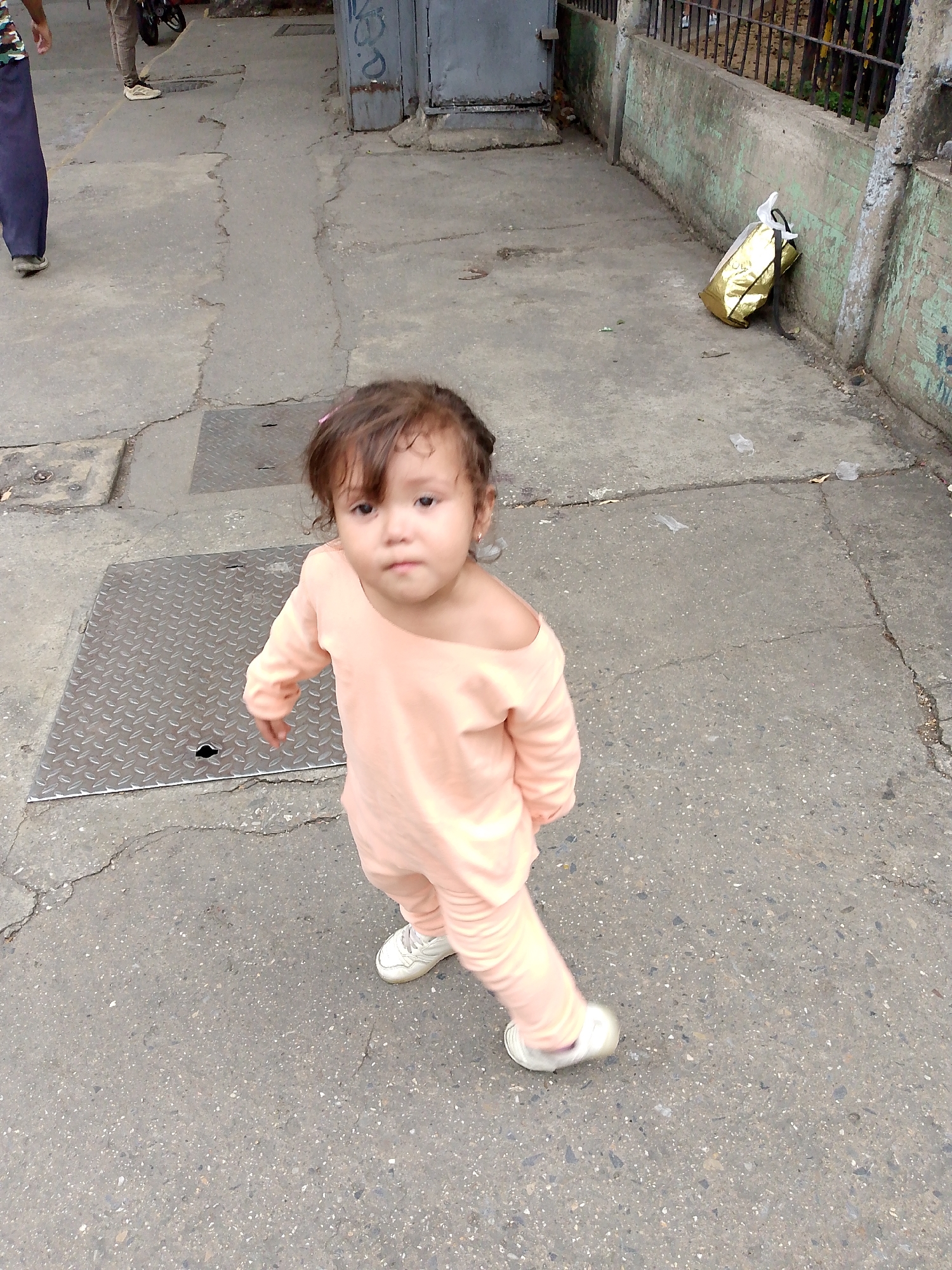 A young child stands on a cracked sidewalk in Caracas, Venezuela. The child wears a light peach-colored outfit and white sneakers. A metal manhole cover is to the child's left. A fence runs along the right side of the image, with a gold shopping bag next to the base of the fence. A door is located on the left side of the image, near the upper portion. Another adult is visible in the upper left corner of the image.