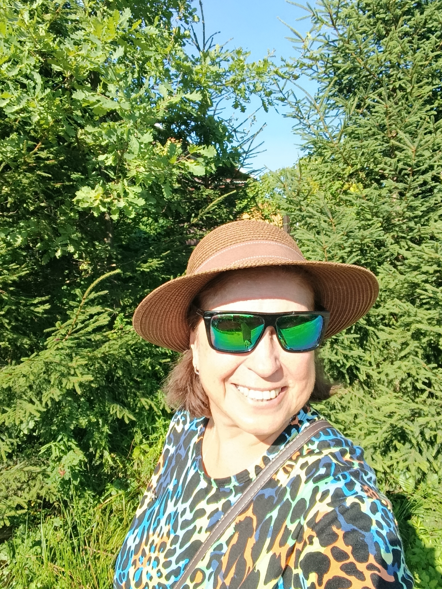 A woman wearing a brown straw hat and sunglasses smiles at the camera in a selfie. She has on a black, blue, orange, and green patterned shirt. The background is filled with lush green trees and a bright blue sky. The lighting suggests it is daytime, likely outdoors. There are no visible landmarks or indications of the specific location within the city of Верея, Russia.