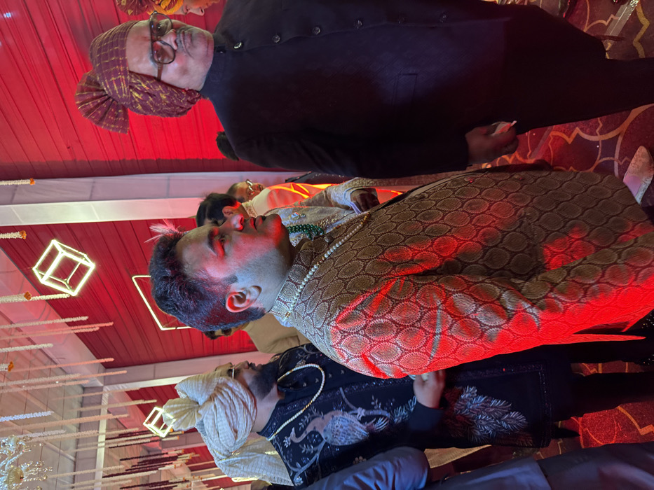 Three men are formally dressed. One wears a dark suit and a turban. The man in the middle has a patterned top with a beaded necklace. The other man wears a dark patterned top and a white turban, adorned with a beaded necklace. The setting seems festive, with red and white decor and cube-shaped lights hanging from above. The photograph has an unnatural rotation to it.
