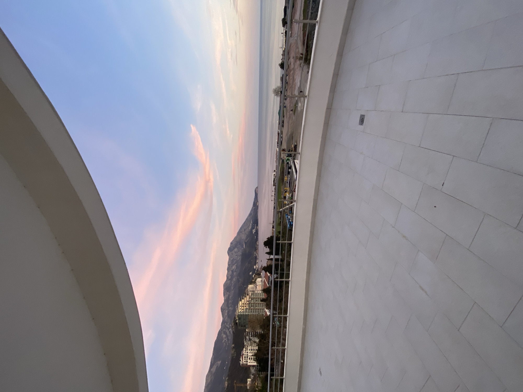 The image presents a panoramic view from an elevated outdoor terrace in Boreti, Montenegro. The immediate foreground shows a light-colored, possibly off-white or beige, tiled floor composed of rectangular tiles laid in a stretcher bond pattern, with a small circular drain visible on the right. A modern railing, constructed from metal uprights and a top rail with glass infill panels, separates the terrace from the vista. Above, a curved, light-toned concrete or stucco overhang partially frames the sky.
Beyond the railing, the scene unfolds into a coastal landscape. To the left, a series of multi-story buildings, likely hotels or residential complexes, are nestled against the base of large, dark mountains. A construction crane is visible among these structures. The central and right portions of the midground reveal a wide beach area, characterized by exposed sand and earth, suggesting either natural topography or ongoing development. A solitary, indistinct human figure is visible on the beach towards the right side.
The calm, light blue-grey water of the Adriatic Sea extends from the beach to the horizon. The sky is a prominent feature, displaying a soft gradient from light blue to a pale pink, with wispy pink clouds streaking across, indicative of either dawn or dusk. A chain of rugged, dark mountains forms the distant background, extending along the coastline to the left. No specific interactions are visible beyond the single pedestrian.