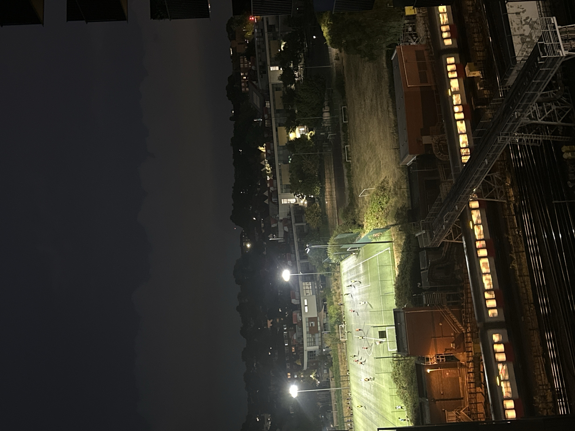 This is a high-angle, nighttime aerial view of a well-lit, artificial turf soccer field in the city of Greater London, United Kingdom. Approximately ten to fifteen individuals, likely players, are visible on the field, engaged in a soccer game under the illumination of floodlights. Several buildings, including what appears to be a modern educational facility with large windows, are situated in the background behind the soccer field. Beyond these buildings, a residential area with scattered lights is visible, set against a dark, overcast sky. In the foreground and lower half of the image, train tracks run horizontally, with a red and white passenger train illuminated by its internal lights, actively moving along the tracks. A pedestrian overpass with metal railings and stairs is also present, spanning across the train tracks. The environment suggests an urban or suburban setting with sports facilities and transportation infrastructure.