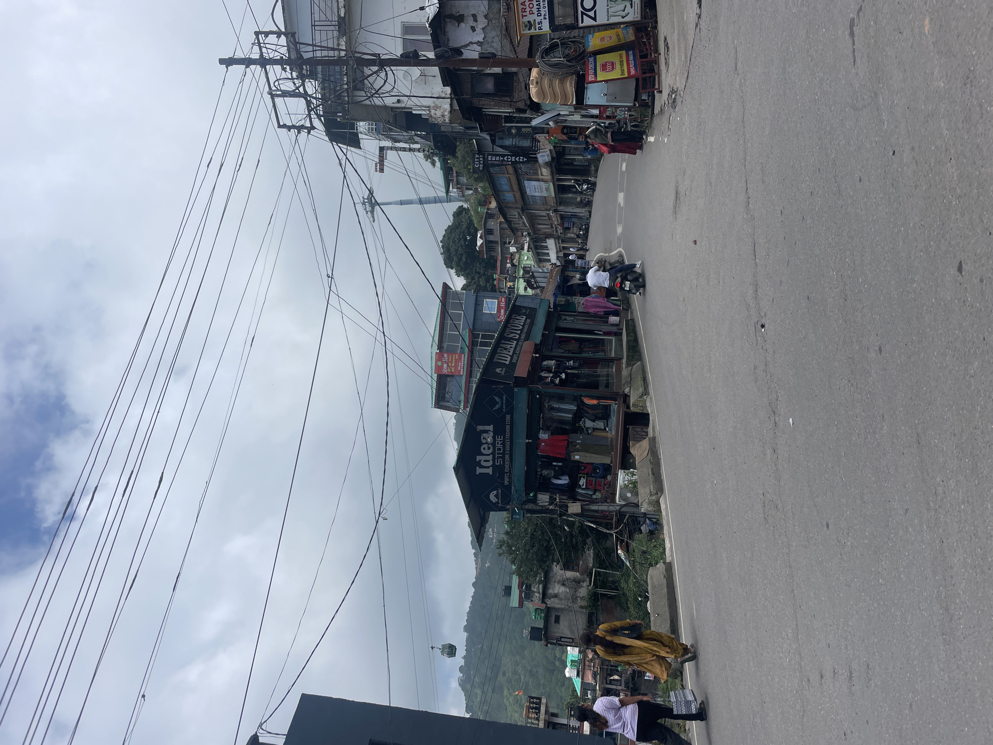 The image depicts a street scene in Dharamshala, India, under an overcast sky. A paved road slopes gently upwards, lined on both sides by buildings and shops. Electrical wires are strung overhead, supported by utility poles.
In the foreground, two women are walking away from the camera on the left side of the road. One is wearing a striped shirt and dark pants, carrying a bag. The other is wearing a mustard-colored dress.
To the right of the road, a shop labeled "Ideal Store" is visible, displaying clothing racks with various garments. Further up the street, a motorbike with a rider is moving away from the camera. Several other people can be seen on the sidewalks and in the distance.
On the left side of the image, a cable car gondola is suspended in the air against a backdrop of lush green hills. The architecture of the buildings suggests a hillside town setting. Signs in English and possibly other Indian languages are visible on the shops, indicating commercial activity. A water tank is mounted on the exterior of a building on the right. There are several advertisements and informational signs on the right side of the road, including one that appears to be from the Traffic Police.
