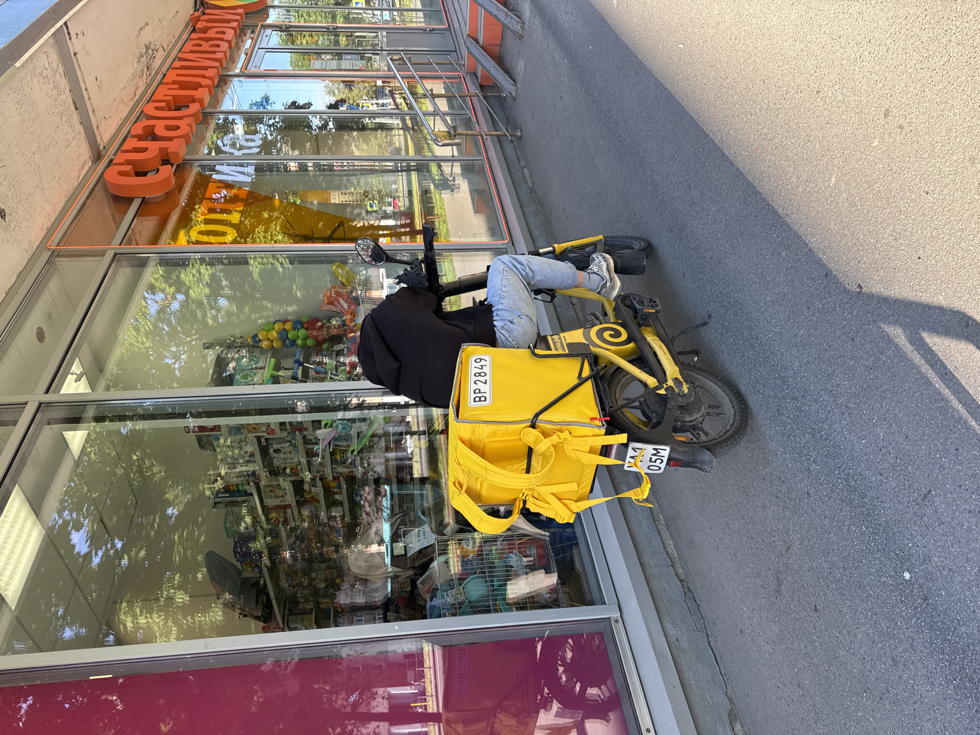 A person rides a yellow electric scooter with a large yellow delivery bag on the back, traveling along a sidewalk next to a store with a bright orange awning and signage. The store's windows reflect trees and the sky. The scooter has a license plate that reads "BP 2849" and below that "YA 1 05M". The person is wearing a black hooded sweatshirt and blue jeans. The scooter is parked in front of the store and the person is on the scooter. The scene appears to be in a city, with buildings and a sidewalk visible. The lighting suggests it is daytime.