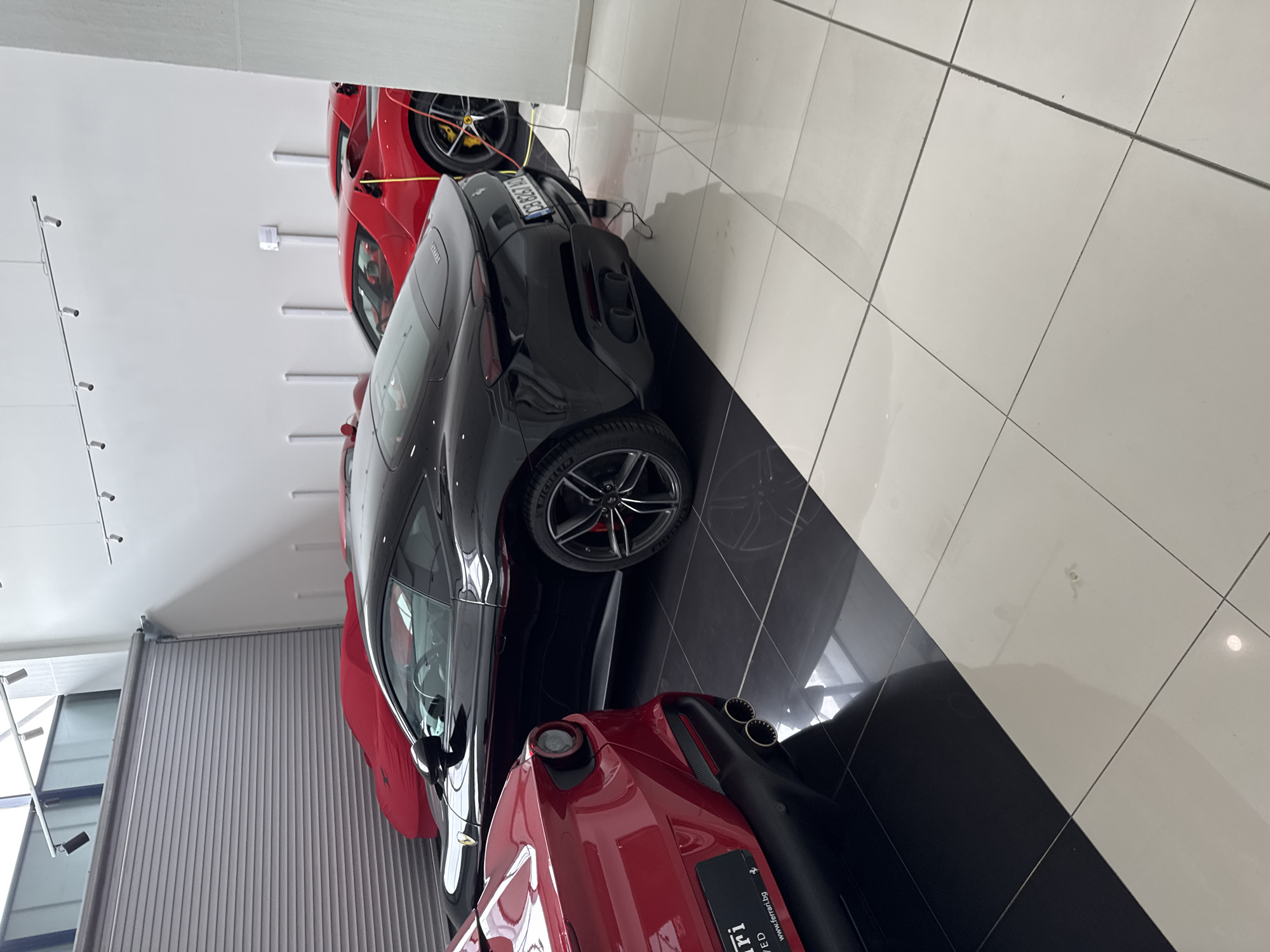 The image shows a showroom with three luxury sports cars. The cars are parked on a black and white tiled floor. There is a red Ferrari at the back, a black car in the middle, and another red Ferrari at the front. The license plate of the black car appears to read "CZ 7828 GT". A roller shutter door and modern lighting are visible in the background. The image was taken inside a building in Sofia, Bulgaria.