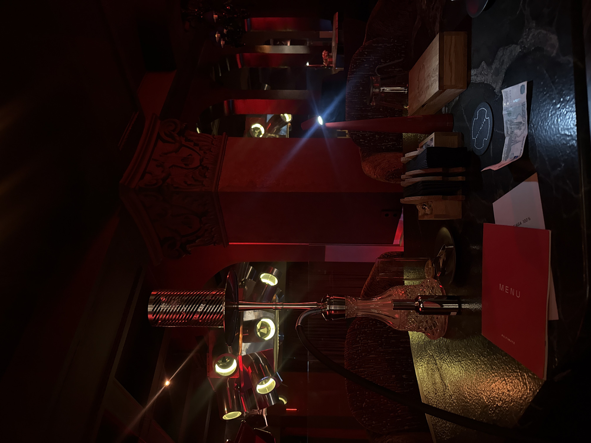 The image depicts the interior of a dimly lit, upscale establishment, possibly a hookah lounge or bar, characterized by rich red ambient lighting and ornate decor. A prominent hookah with a metallic shaft and a glass base stands on a textured dark table in the foreground. To the left, a unique light fixture with multiple spotlights illuminates the scene, casting a dramatic glow.
The table is also adorned with a red menu, a small glass, a metallic bottle, and a coaster. In the background, plush velvet chairs are visible, suggesting a comfortable seating area. The architecture features decorative columns and arched doorways, contributing to an opulent atmosphere. A slim, red conical lamp with a bright spotlight emanates a beam of light, adding to the dramatic lighting effect.
The overall ambiance is one of luxury and sophistication, with the red lighting creating a warm and intimate setting. The scene is devoid of any visible individuals, allowing the viewer to focus on the details of the environment and the objects within it. The presence of a menu and the type of establishment suggest a place for socializing and relaxation.