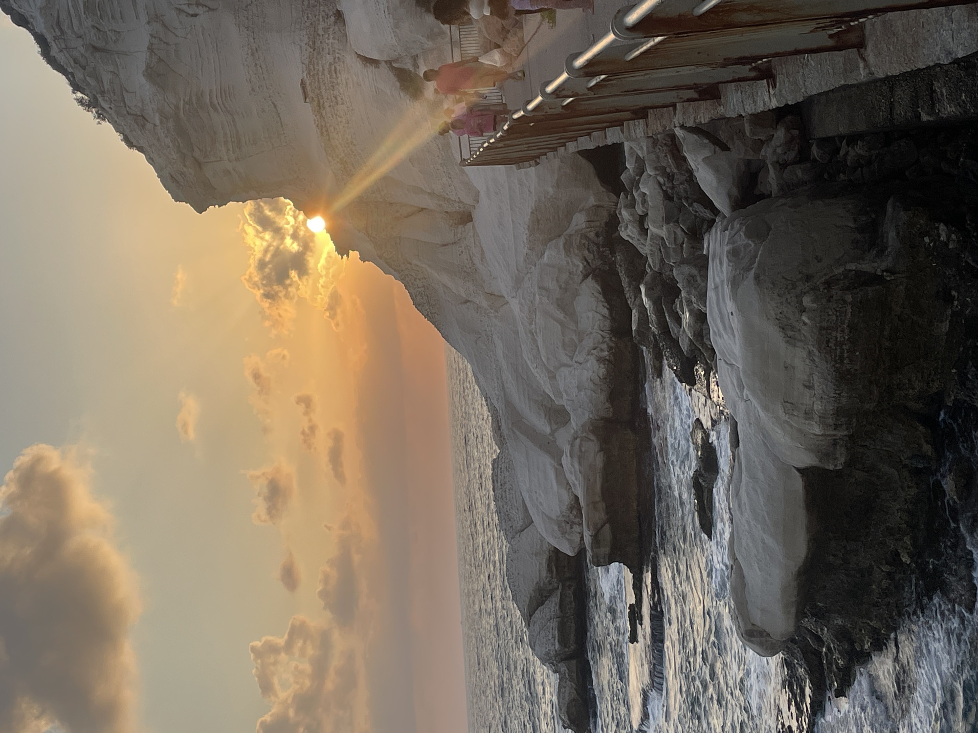 This image captures a sunset over the Mediterranean Sea at Rosh HaNikra in the Mate Asher Regional Council, Israel. The sun, partially obscured by a white cliff face, casts a warm orange glow across the sky and reflects off the water. The cliff, a prominent geological feature, is layered and appears to be made of chalk or limestone. A pathway, bordered by a weathered metal railing, runs along the cliff's edge, and several individuals, including a person in a red shirt carrying a white bag and a person in a pink jacket, are walking on it. The sea is choppy, with waves breaking against a rocky shoreline composed of large, light-colored boulders and dark, jagged rocks closer to the water. The scene is a natural coastal landscape enhanced by a man-made pathway, suggesting a tourist or recreational area.
