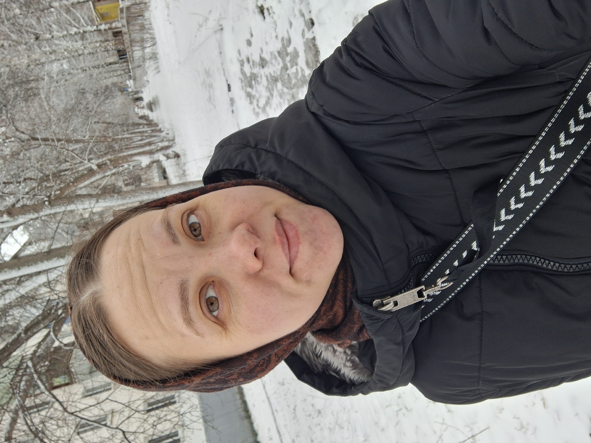 A woman is posing for a selfie outside, likely in Chaikovsky, Russia, based on the background surroundings which include multi-story residential buildings, snowy ground, and bare trees. The woman is wearing a dark, puffy winter jacket and a patterned headscarf. The jacket features a visible zipper and a shoulder strap with a geometric design. Her face is prominent in the foreground, and she appears to be smiling slightly. The background shows a snow-covered area with trees and buildings, providing context to the location and the winter season.