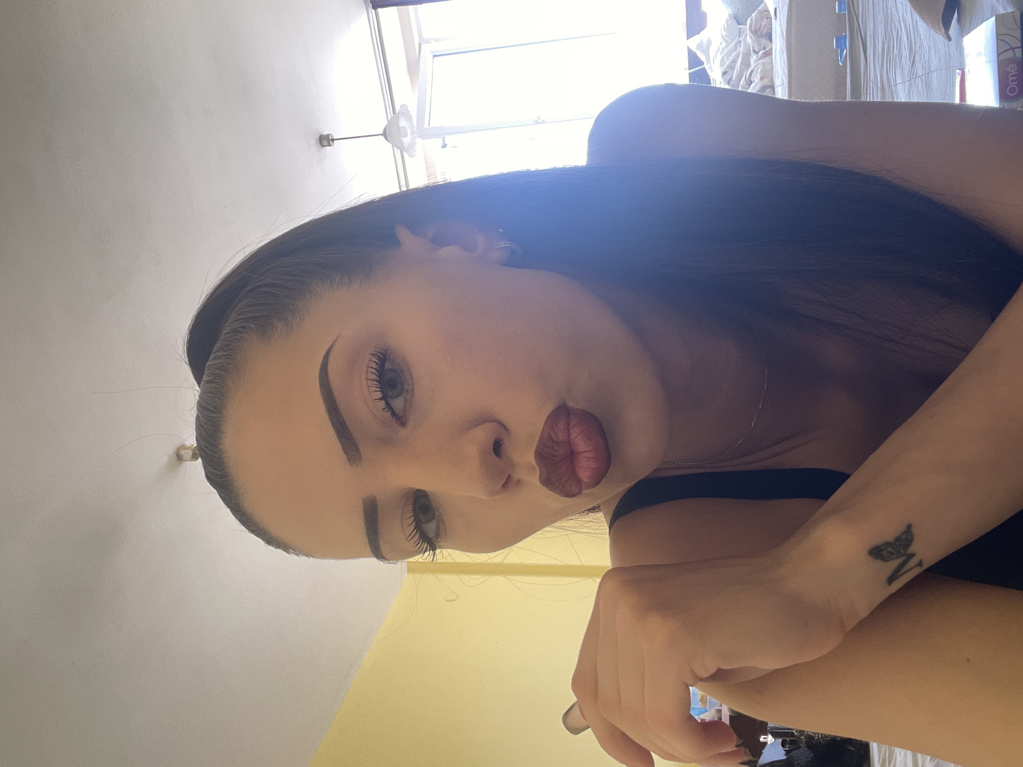 The image displays a head-and-shoulders portrait of a young woman, taken from a slightly low angle, as if looking up at her. She has long, dark hair that sweeps over her right shoulder and cascades down her back, mostly out of frame. Her facial features include light-colored eyes, prominent dark eyebrows, and full lips pursed into a "kiss face" or pout, accentuated by dark red lipstick. She wears a delicate silver chain necklace and a dark-colored top, possibly a tank top or camisole.
Her left arm is visible in the lower right portion of the frame, bent at the elbow. A small tattoo, appearing to be a stylized letter "N" with wings, is visible on her left wrist.
The setting appears to be an indoor room during the daytime, indicated by bright natural light streaming in from a window in the upper background. A white wall or ceiling takes up a significant portion of the background, with a light fixture (possibly a ceiling fan or lamp) partially visible near the top left. In the upper right background, indistinct white and light-colored fabrics or bedding suggest a bedroom. A white box with the partial text "Ome" in red is also visible in the background on the far right. The overall impression is one of a casual, playful pose in a well-lit indoor environment.