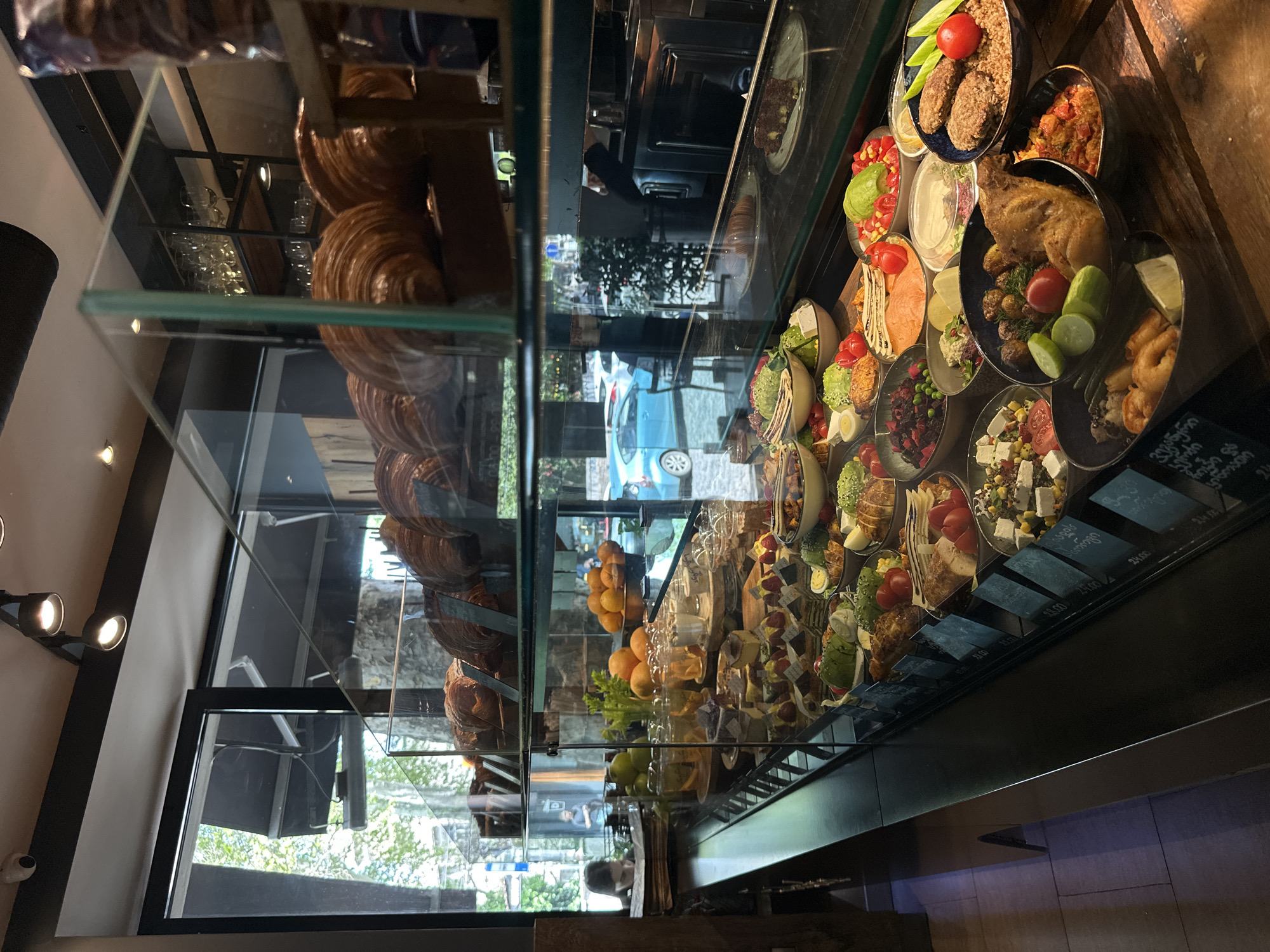 The image depicts the interior of a cafe or deli, focusing on a brightly lit glass display case filled with a variety of food items. The perspective is angled, looking through the display towards the outside street, creating reflections and transparent views. The overall image is rotated 90 degrees clockwise.
On the left side of the display, large quantities of golden-brown croissants or similar baked goods are stacked on shelves. The right side of the case showcases numerous individual portions of diverse dishes presented in bowls and containers. These include various salads with fresh vegetables like cherry tomatoes, lettuce, cucumber, and ingredients such as eggs, grilled chicken, salmon, and feta cheese. Other savory options feature meatballs or patties, roasted chicken with potatoes, shrimp, and what appears to be grain-based dishes. There are also fruit salads with berries and melon, and small dessert items. Oranges and other whole fruits are also visible on a middle shelf.
Through the glass, a street scene is visible with a light blue car and trees, indicating a daytime setting in an urban environment. Reflections on the glass show elements of the cafe's interior, including ceiling lights and faint outlines of people, suggesting activity within the establishment. Beyond the display, empty glasses are stacked on shelves.
Below the individual food portions on the right, dark placards with white text are visible. Some clearly display prices, such as "23.00" and "24.00". Other placards contain text written in Georgian script, such as "სალათი" (salati), which means "salad," further confirming the location context of Tbilisi, Georgia. The cafe appears to offer a wide selection of freshly prepared meals and pastries.