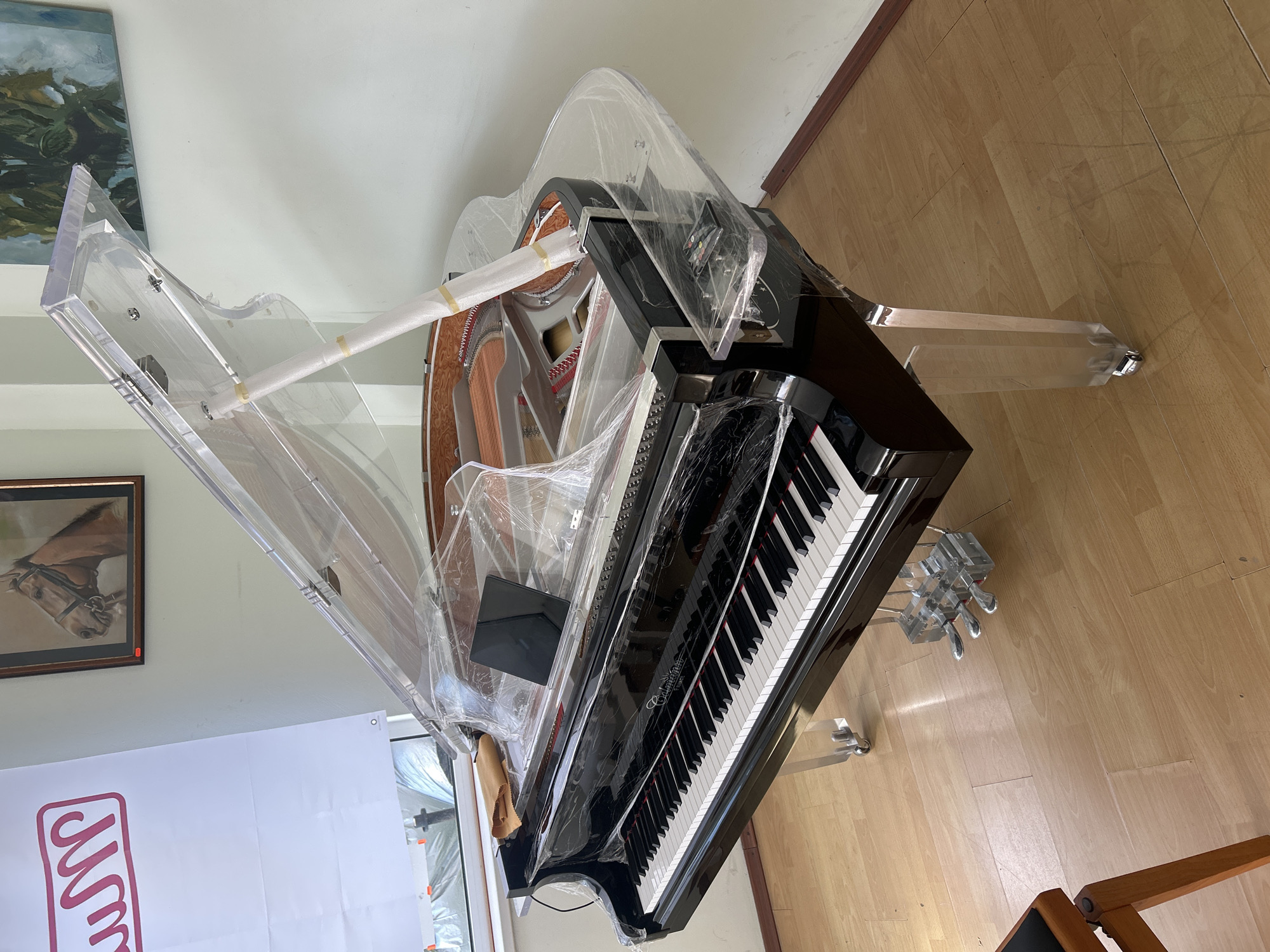 The image shows a grand piano, possibly a display or showroom model, in a room with light-colored walls and a wood floor. The piano is predominantly black with white keys and appears to have some clear acrylic or plastic elements in its construction, including the legs and some parts of the case. It is partially covered in protective plastic wrapping. The piano is surrounded by other objects including part of a banner with red text, what appears to be a framed picture with a horse, and furniture. The location is potentially a commercial space, possibly in Plovdiv, Bulgaria. There are no people visible.