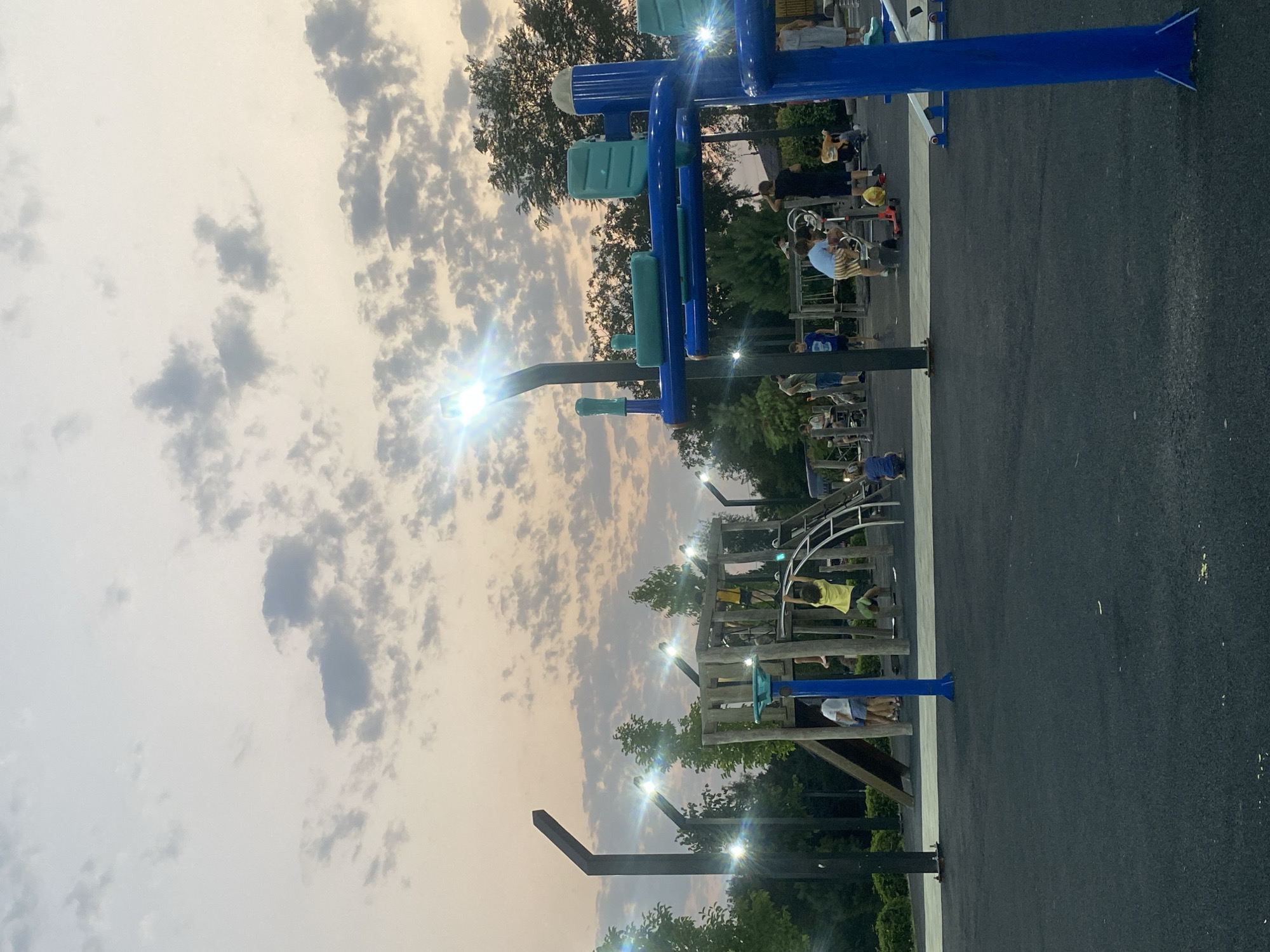 This image captures an outdoor playground in Lagodekhi, Georgia, during twilight. The sky is a soft gradient of pale orange and grey, filled with scattered, textured clouds. Several tall, modern lampposts with integrated lights illuminate the scene, casting bright, starburst-like glares. The playground itself features a mix of wooden and metal structures. A wooden playhouse with a slide is visible on the left, with several children on or near it. To the right, a blue and green metal exercise machine stands prominently.
Various individuals, including adults and children, are scattered throughout the playground. Some children are actively playing on the equipment, such as a child hanging from monkey bars on the wooden structure, and another on a scooter. Others appear to be socializing or observing. The ground is a dark, rubberized surface typical of modern playgrounds, with lighter grey sections marking pathways. Lush green trees and foliage form a backdrop behind the playground structures, suggesting a park or recreational area. The overall atmosphere is calm and family-friendly, despite the artificial lighting indicating the approaching evening.