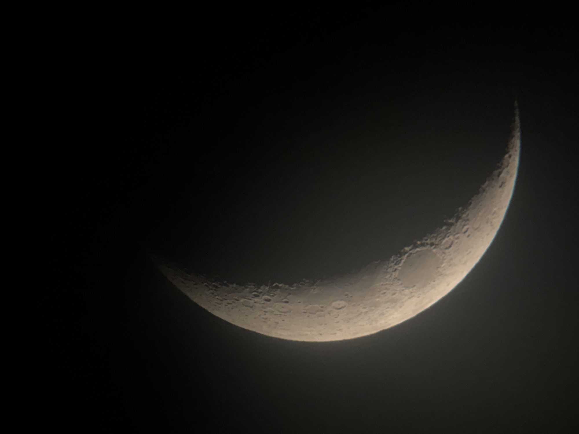 The image shows a crescent moon against a dark sky. The moon is illuminated on its right side, showing craters and surface features. The background is black, indicating nighttime. There are no people or other objects visible in the image. The scene is a close-up view of the moon, likely taken with a telescope or a camera with a powerful zoom lens. There are no visible text elements. The overall mood is serene and cosmic.