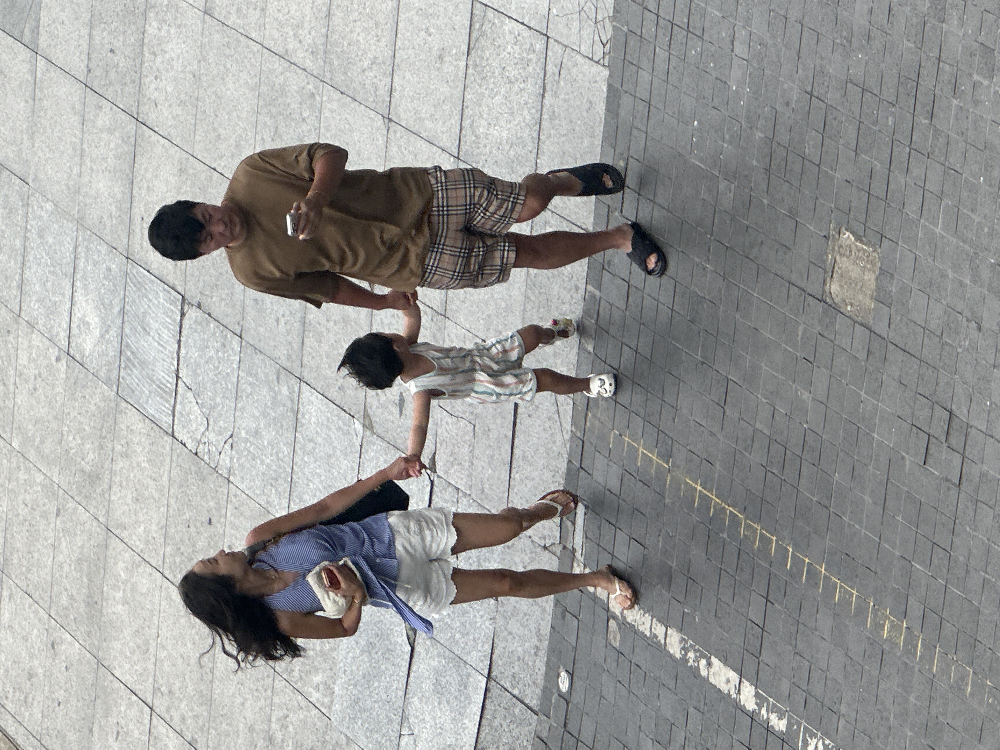 A family, composed of a woman, a man, and a child, walks across a paved city square in Đà Nẵng, Vietnam. The woman, on the left, wears a blue and white striped sleeveless top and white shorts. She holds a small, light-colored clutch in her left hand and the child's right hand in her left. The man, on the right, is dressed in a brown t-shirt and plaid shorts. He holds a smartphone in his right hand and the child's left hand in his left. The child, positioned between them, wears a striped outfit. They are all wearing sandals. The ground is paved with square tiles of varying shades of gray. A white painted line and a series of yellow dashes are visible on the pavement, suggesting a crosswalk or pedestrian marking. The lighting suggests it is daytime, with shadows cast by the individuals. The overall scene depicts a casual stroll through a public urban space.