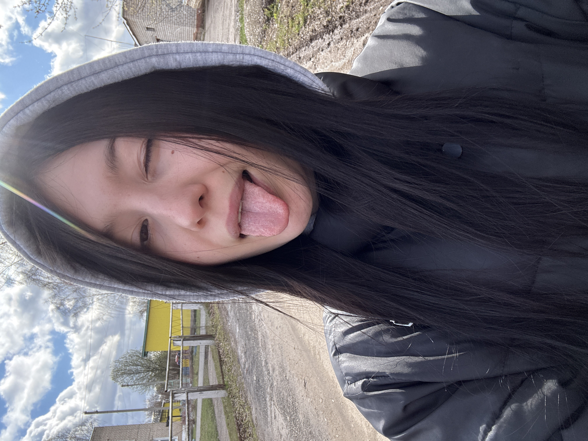 A playful outdoor selfie features a young person, likely female, with long, dark hair, wearing a grey hooded sweatshirt with the hood up and a dark puffer jacket. Her head is tilted, and she looks towards the camera with one eye partially closed, playfully sticking out her tongue. A bright sun flare with a rainbow effect is visible on the left side of the image, suggesting sunny weather.
The background reveals a rural or semi-rural setting, consistent with Фоки, Russia. The sky is bright blue with scattered white clouds. A dirt or gravel road stretches behind the subject, bordered by sparse vegetation, possibly early spring trees and bushes. In the distance, simple structures are visible, including a brick house and a yellow building, separated by fences. The scene is illuminated by broad daylight, capturing a casual and lighthearted moment. There is no visible text.