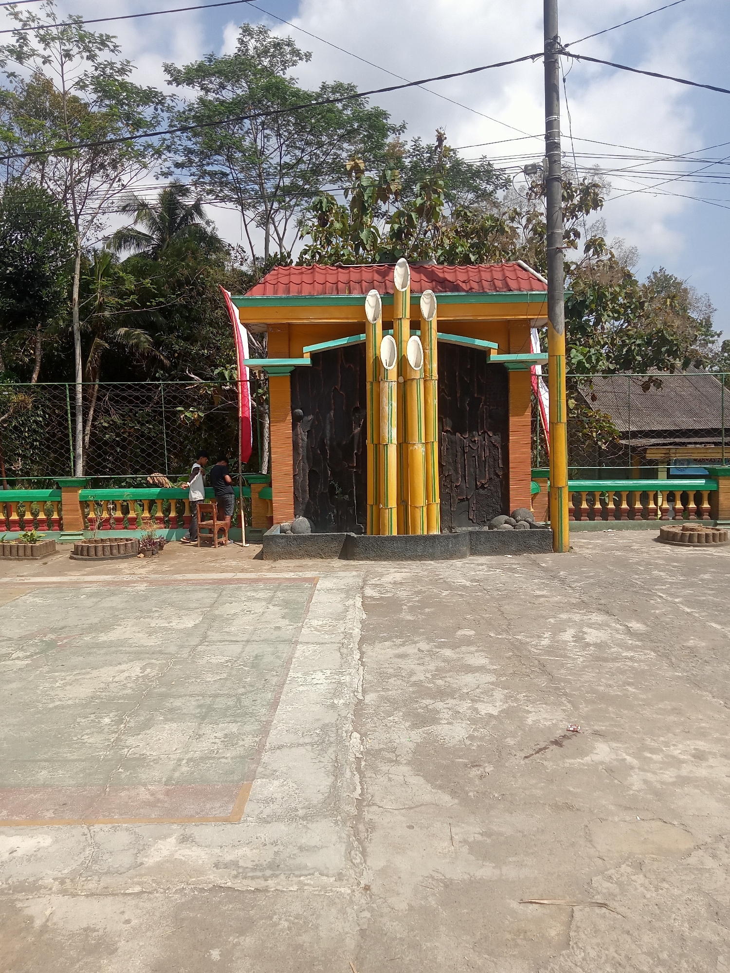 The image depicts a small, colorful monument with a red-tiled roof and yellow walls, featuring decorative bamboo-like structures and dark, textured panels, situated outdoors in Pule, Indonesia. In front of the monument is a concrete paved area, possibly a plaza or sports court, with faded markings. To the left of the monument, two young males are present near a green and yellow fence. One is wearing a white t-shirt and dark shorts, standing next to a wooden chair. The other, in a black t-shirt and shorts, appears to be looking at something on the ground. Behind the fence, dense foliage and trees are visible, with a cloudy sky overhead. A utility pole with electrical wires stands to the right of the monument. The environment appears to be somewhat unkempt, with some litter visible on the concrete.