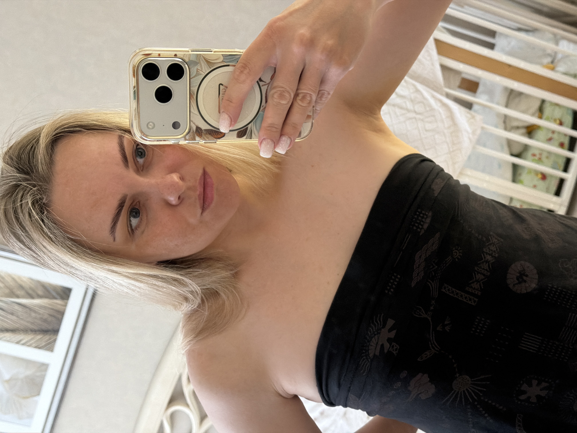 The image is a brightly lit, vertically oriented selfie taken by a woman indoors. A blonde woman with light-colored eyes and fair skin looks directly at the camera with a neutral expression. She is captured from the chest up, wearing a dark, strapless top or dress featuring subtle, intricate patterns that appear to be tribal or nature-inspired.
Her right hand, adorned with nails manicured in a white glitter ombre style, holds up an iPhone. The phone is encased in a clear cover with a delicate floral design, and a pop socket or ring holder on its back displays what might be part of the word "Venti." The reflection in the phone clearly shows its three camera lenses.
The background reveals a home interior. To the woman's right, a white wooden crib with white and green bedding is partially visible, indicating a nursery or bedroom setting. The soft, even lighting suggests it is daytime indoors. This personal moment was captured in a domestic environment in Votkinsk, Russia.