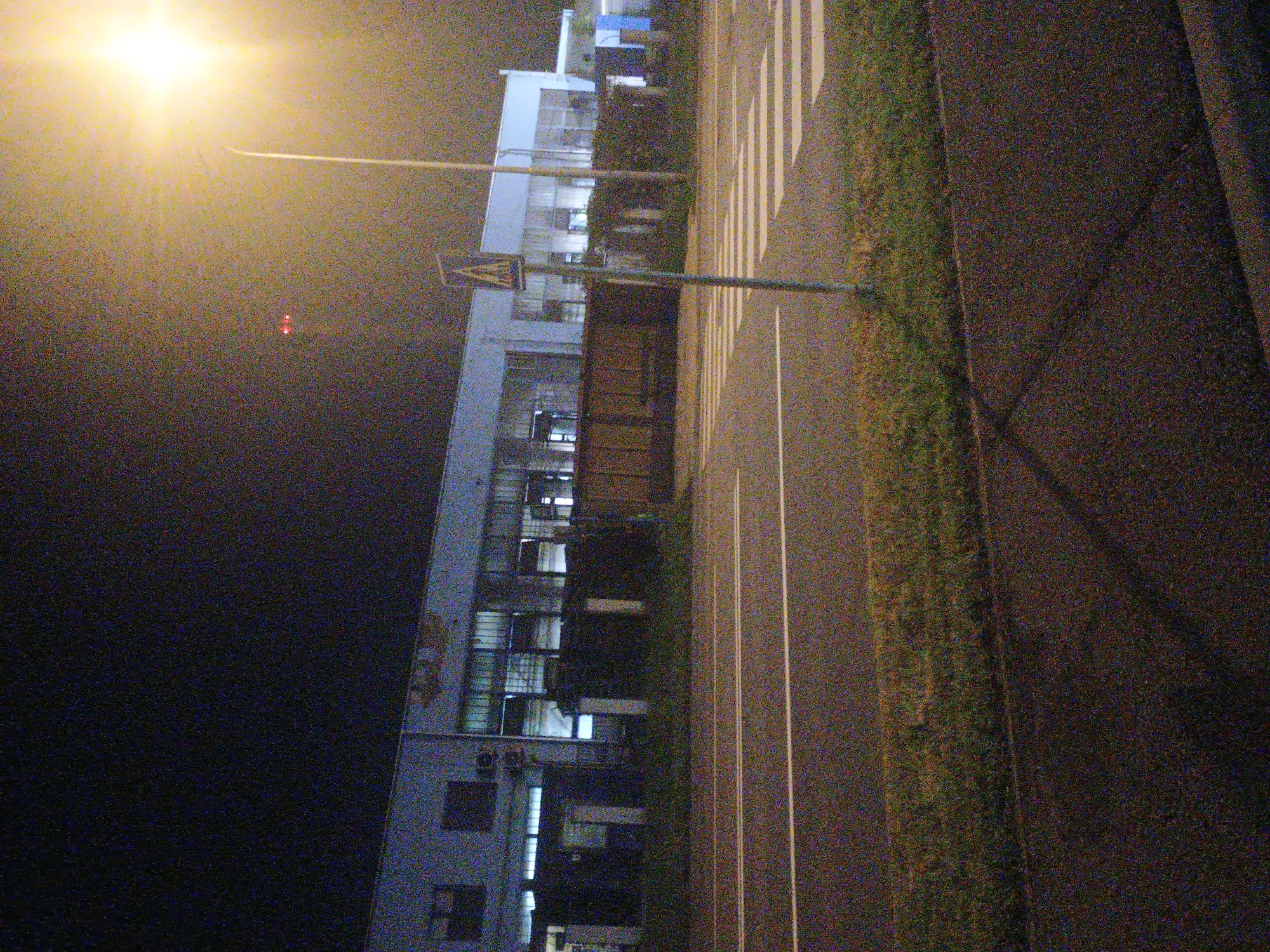 This is a nighttime photograph depicting a street scene in an urban or industrial area, likely in Kroměříž, Czechia. The image exhibits significant digital noise, characteristic of low-light photography.
To the left, a multi-story building with a light-colored facade stretches vertically, featuring numerous windows, some of which show illuminated interiors. The building appears to be commercial or industrial rather than residential.
Running through the middle of the frame is a paved road. It is marked with a distinct white pedestrian crossing (zebra stripes) and additional solid white lines, possibly delineating lanes or the road's edge. A tall pole stands by the road, supporting a diamond-shaped pedestrian crossing sign. Another pole extends from the bright light source, suggesting a street light. Adjacent to the road, a strip of grass or weeds is visible. The foreground and the right side of the image are dominated by dark pavement.
The scene is primarily lit by a very bright street light in the upper left, casting a strong glow and creating lens flare or atmospheric haze in the dark sky. The rest of the scene is dimly lit, emphasizing the nighttime setting. In the dark background above the building, a few small red lights are faintly discernible. No discernible human activity or specific event is taking place, contributing to a quiet, deserted atmosphere. No explicit text is visible.