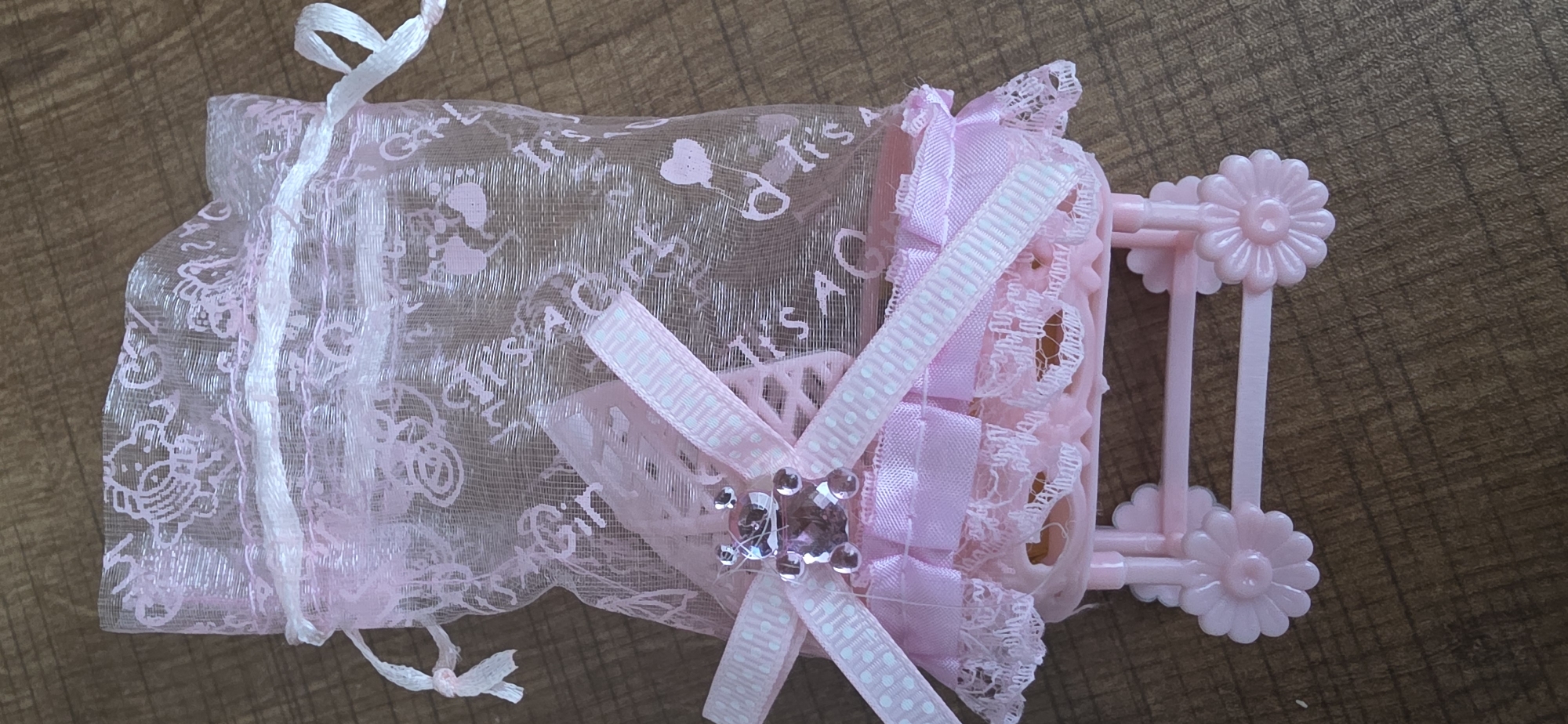 A pink, sheer organza gift bag with a white drawstring, adorned with faint pink baby-themed prints (e.g., rattles, safety pins, hearts, and text), rests on a brown wooden surface. The bag is partially open at the top and cinched closed with a white ribbon. Below the bag, a miniature pink plastic bassinet, decorated with pink lace trim and ribbons, sits on a light pink plastic frame with four flower-shaped wheels. A pink ribbon with white polka dots is tied in a bow on the front of the bassinet, centered by a small, clear and pink rhinestone-encrusted teddy bear charm. The scene is photographed from a slightly elevated, close-up perspective. There is no indication of the city of Sumgayit, Azerbaijan.
