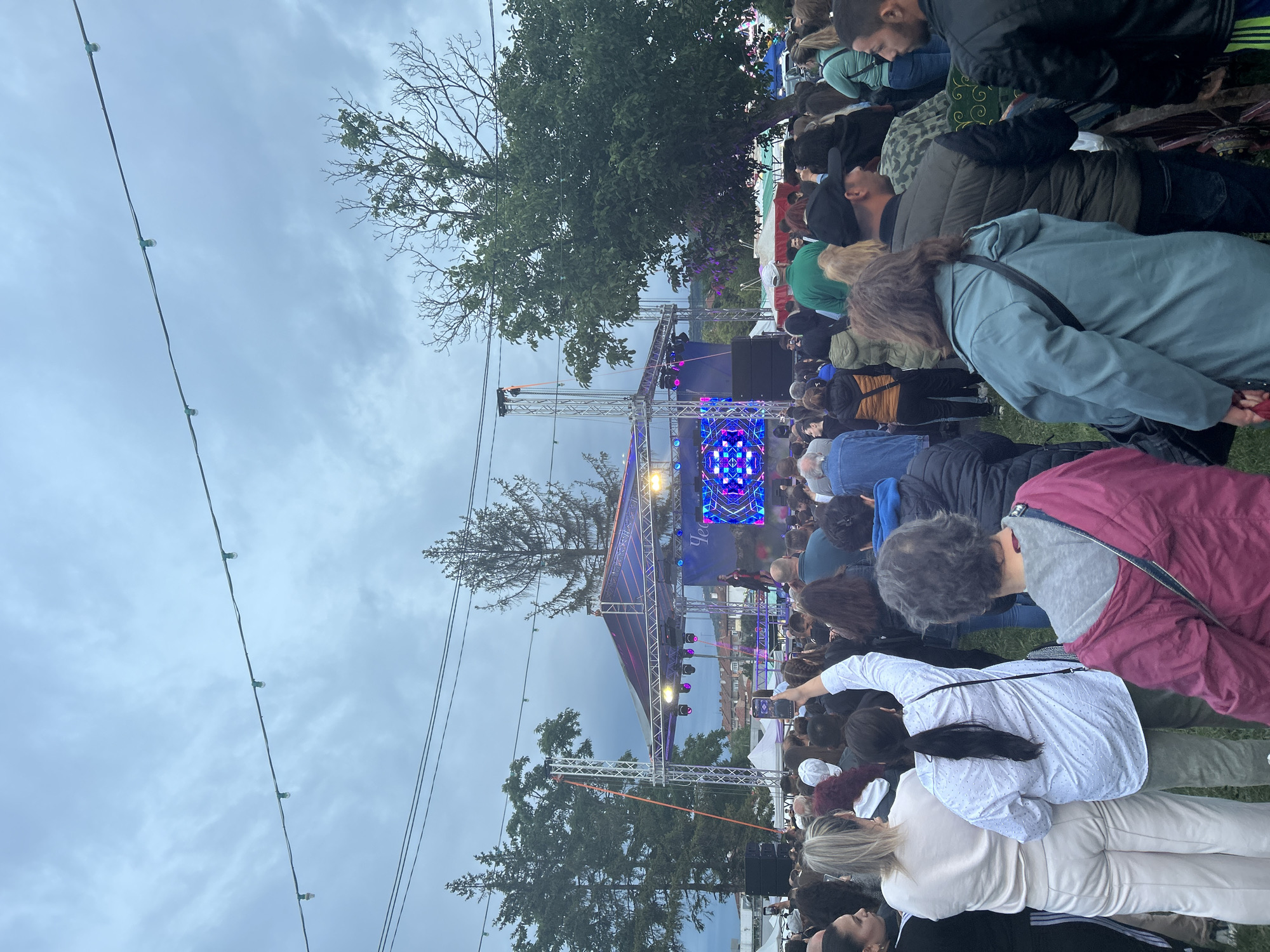 An outdoor concert is taking place in the city of Gorna Oryahovitsa, Bulgaria. A crowd of people is gathered in front of a stage that has a large screen displaying a colorful, geometric pattern. There are lights on the stage and some mist or smoke is present. String lights are visible overhead against a cloudy sky. Trees and buildings can be seen in the background. The attendees are facing the stage, with many holding up their phones to record the event.