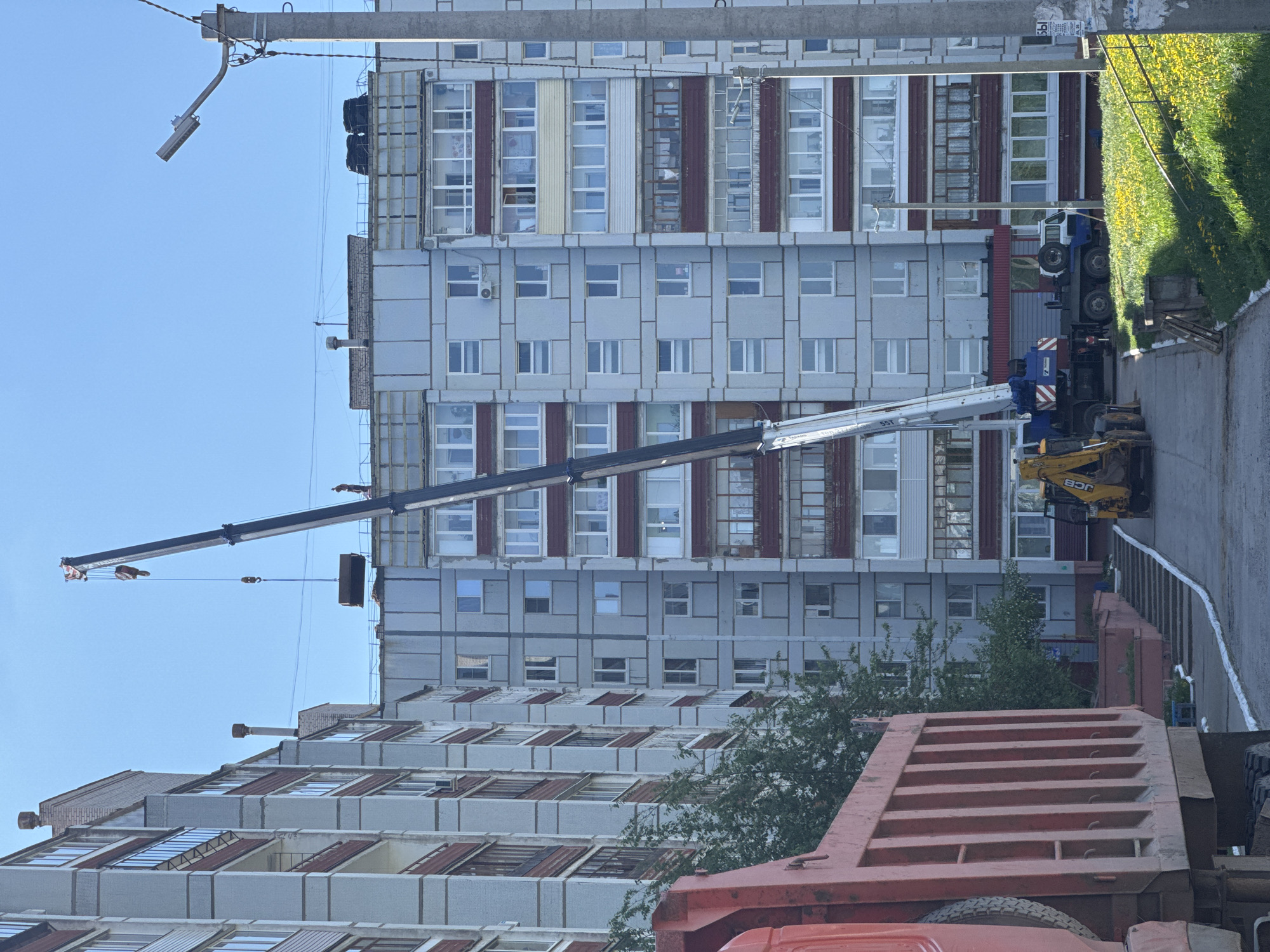 The image shows a construction scene in what appears to be an urban area. A large crane is positioned beside a tall, multi-story building with a repetitive grid-like facade, likely a Soviet-era apartment block. The crane is lifting a rectangular object to the top of the building. A yellow JCB backhoe loader is parked near the crane. A red dump truck is visible in the foreground. Green foliage lines the edge of a nearby walkway. Based on architectural style and available information, this location is most likely the city of Chaikovsky, Russia. There are no people visible in the image.