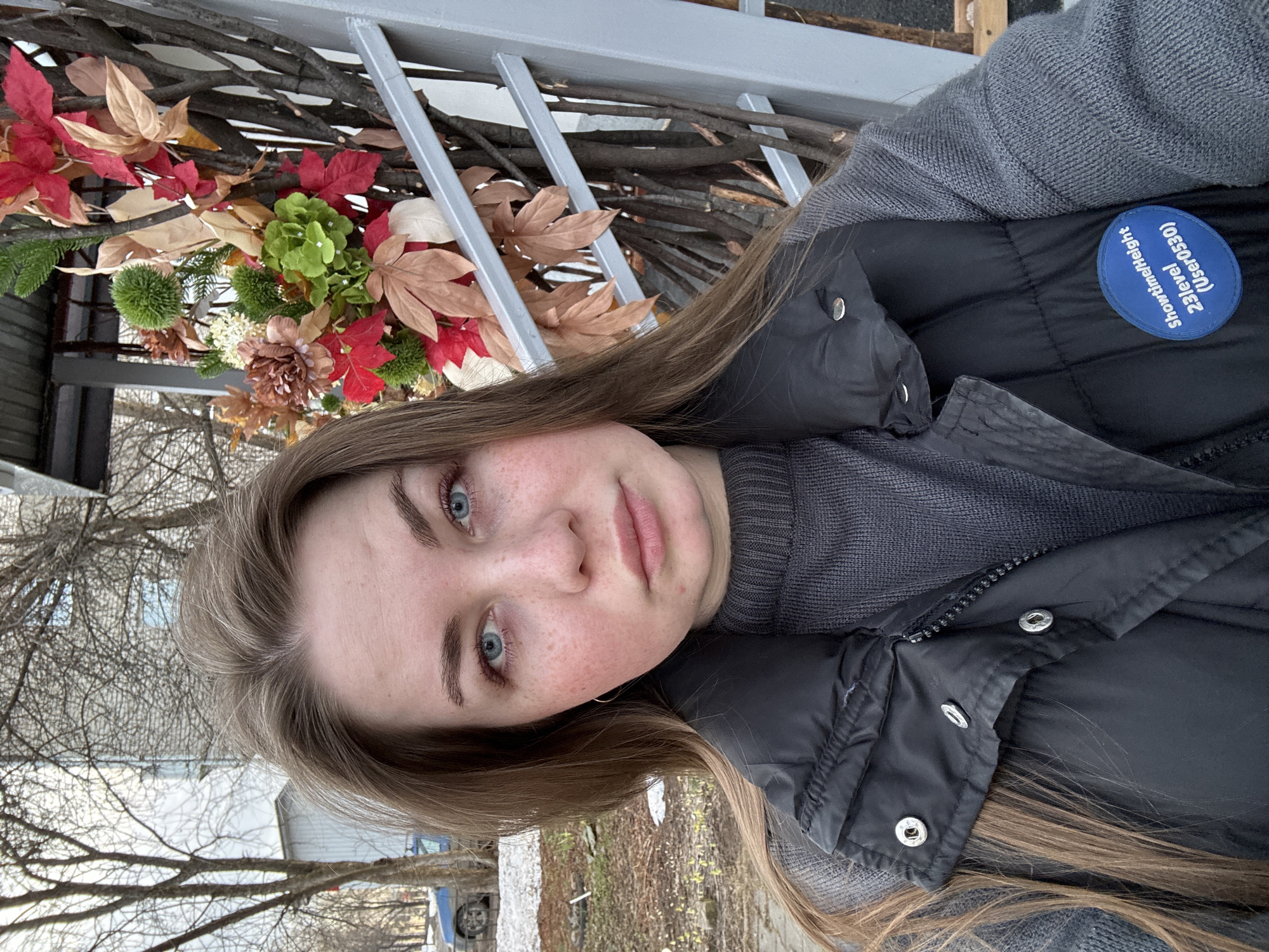 A young adult female is depicted in a close-up, frontal shot, appearing to be a selfie taken outdoors in Chaikovsky, Russia. She has light brown hair, blue eyes, and visible freckles across her nose and cheeks. She is wearing a dark grey ribbed knit sweater underneath a black puffer vest, which is partially unzipped, revealing silver snap buttons on the collar flap. A blue, oval-shaped badge with white text reading "Showtime/Height 23level (User0530)" is affixed to the left side of her vest. Her expression is neutral as she looks directly at the camera.
To her left, the background shows bare deciduous trees, indicative of late autumn or early spring, with a hint of a light-colored building in the distance and a partially visible blue car further back. The ground in this area consists of dirt and patches of melted snow or ice. To her right, a decorative outdoor structure made of intertwined bare branches is visible, adorned with artificial foliage including red, green, and brown leaves, green spherical elements, and floral clusters in various shades. A grey metal railing runs diagonally upwards in front of this decoration. The overall scene suggests an urban or suburban outdoor setting in Chaikovsky, Russia.