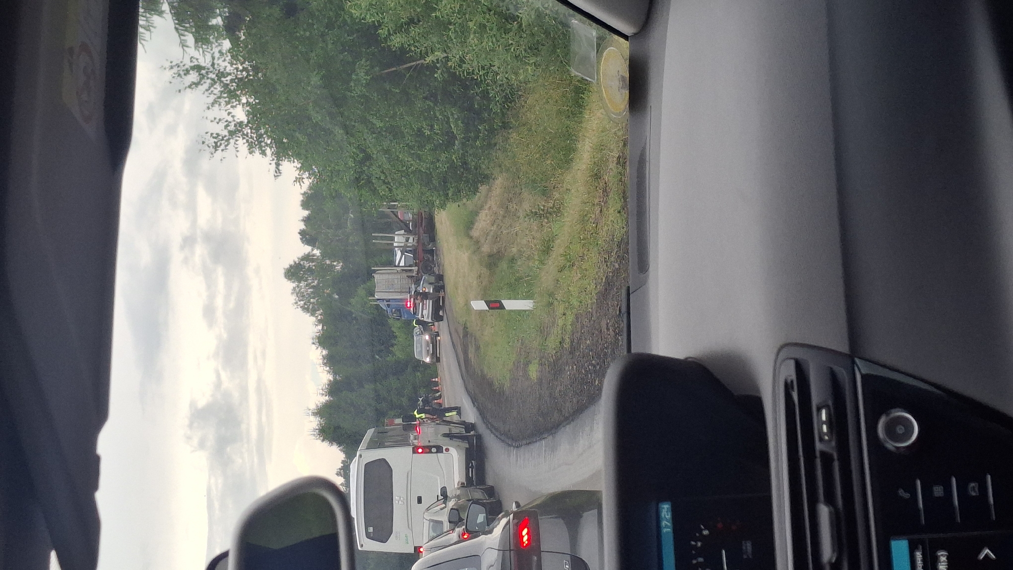 The image depicts a traffic incident on a two-lane road, possibly in the vicinity of Ilyanskiy selskiy Sovet, Belarus, given the provided context. A white bus is stopped on the left side of the road, with its hazard lights illuminated. Adjacent to the bus, two individuals in yellow safety vests appear to be directing traffic or assessing the situation. Further down the road, a convoy of vehicles is stalled, including a large truck with a trailer carrying logs. A police car with flashing blue lights is positioned near the truck, and several other vehicles are visible, suggesting a significant traffic disruption. The road surface is dark and appears wet, with overgrown grass and trees lining the right side of the roadway. The sky is overcast, with a muted, cloudy appearance. The image is taken from the perspective of a vehicle in traffic, looking towards the scene.