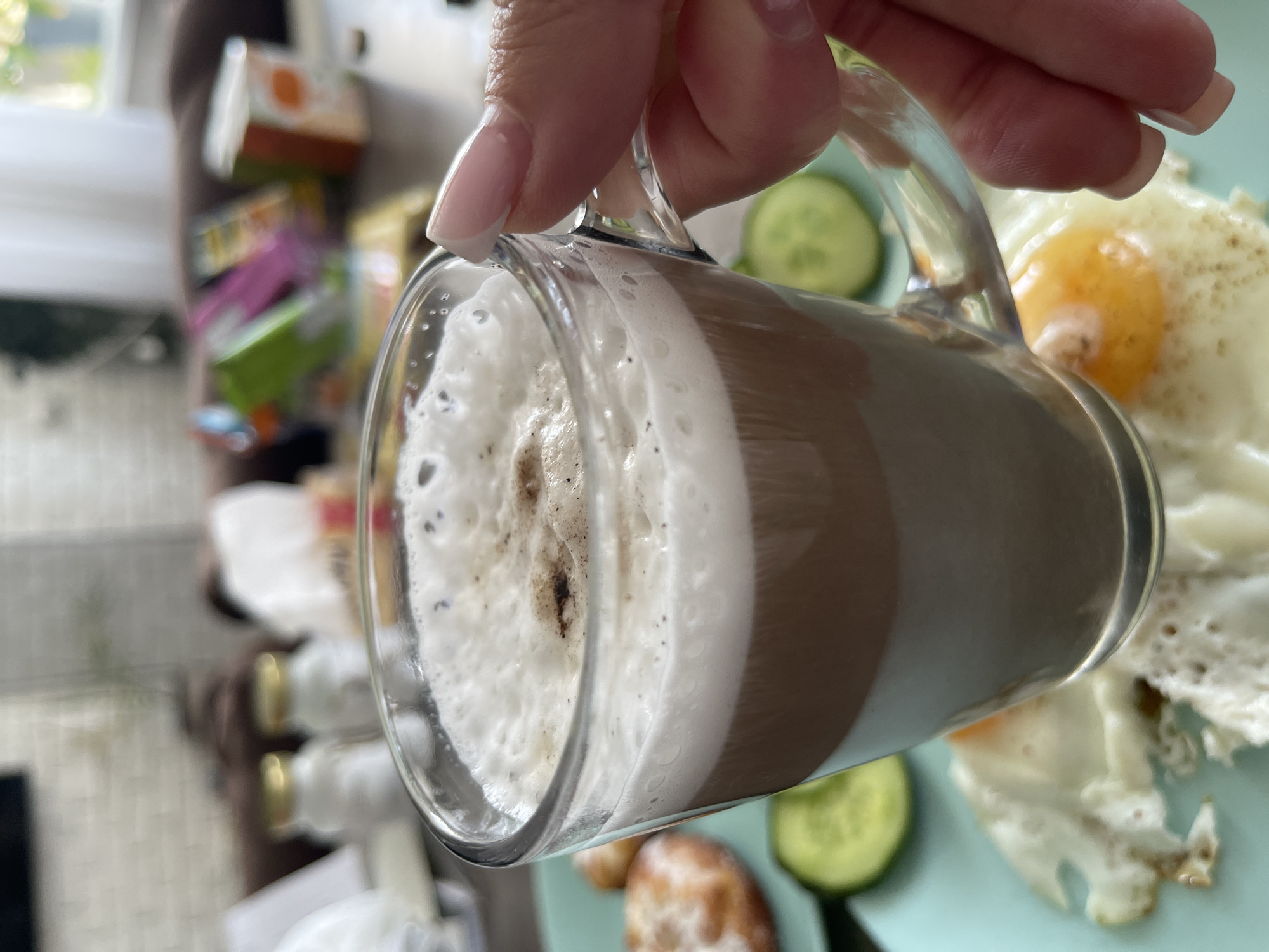 A hand with manicured nails holds a clear glass mug filled with a layered beverage, likely coffee with milk foam, over a plate of breakfast. The breakfast consists of two fried eggs with runny yolks and a few slices of cucumber. In the background, out of focus, are various items including what appear to be small condiment bottles, a white tissue, and brightly colored packaging of snacks. The scene is set indoors, with a blurry background suggesting a domestic setting. The overall impression is of a leisurely morning meal.