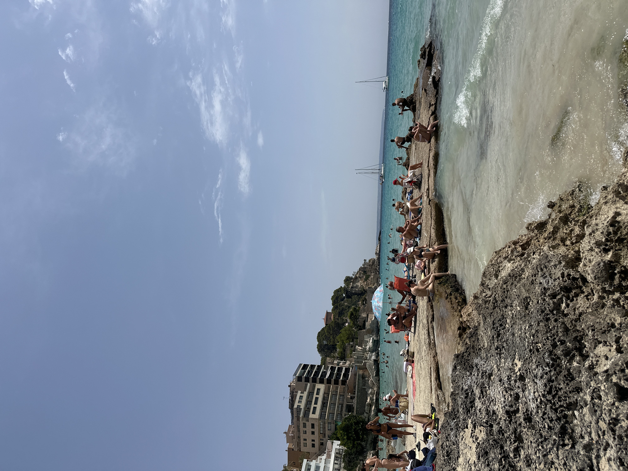 This is an outdoor, eye-level, full shot of a crowded beach scene in Sant Agustí, Spain, on a sunny day with a clear blue sky and a few wispy clouds. In the foreground, rough, dark-grey, porous rocks extend into the clear, shallow, turquoise water, which has small waves lapping at the shore.
Further back, a strip of sand and larger rocks serves as a sunbathing area for many people. They are dressed in swimwear, lounging on towels and chairs, reading, or socializing. A brightly colored umbrella with a blue and white pattern stands on the rocky shore.
Beyond the immediate beach, the clear turquoise water is populated with swimmers and a few boats. In the mid-ground, several white sailboats are visible on the horizon.
To the left of the frame, a multi-story building with a pale facade, likely apartments or hotels, is situated on a hillside overlooking the beach. This building, along with others further down the coast, suggests an urban or semi-urban coastal setting. The overall impression is a busy, picturesque beach resort.