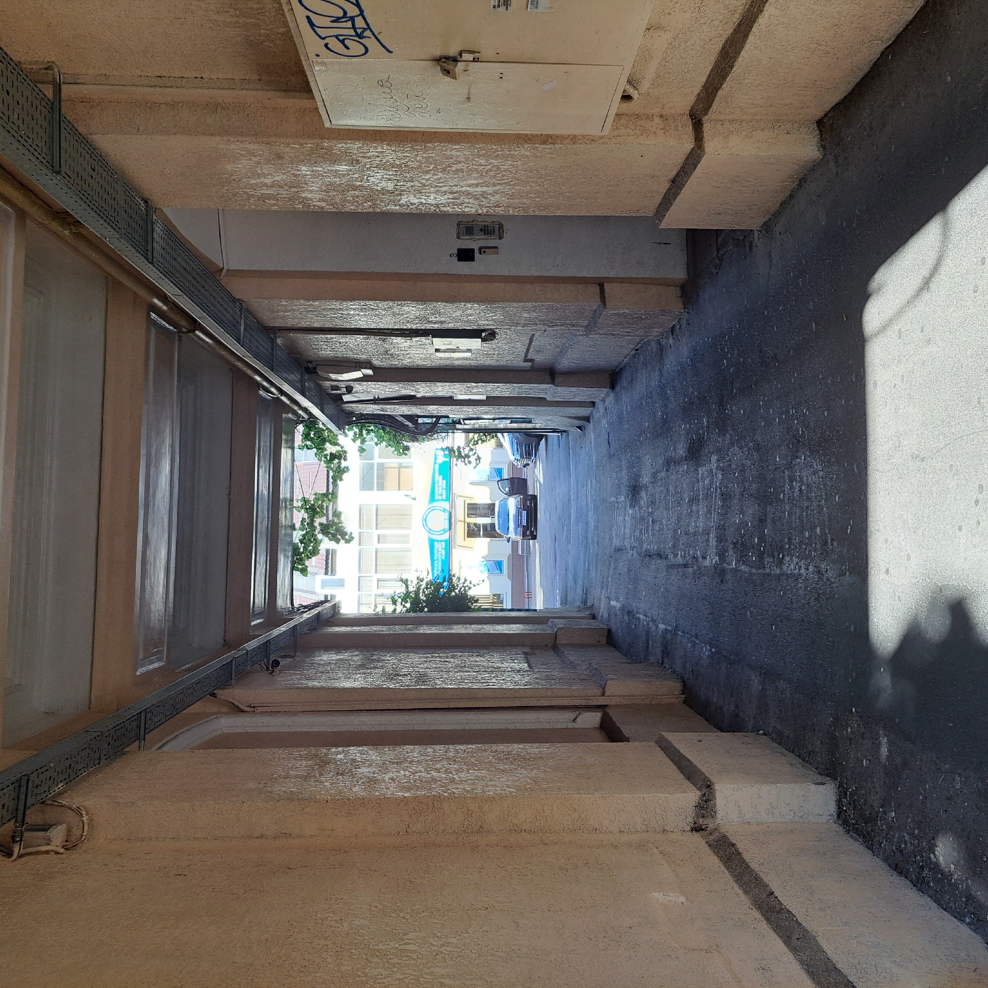 This is an eye-level, medium shot of a narrow, covered alleyway leading to a bright street scene in Odesa, Ukraine. The alley walls are constructed of textured, light-colored concrete or plaster. Electrical conduits run along the ceiling of the alley, supported by metal trays. On the right wall, there is a beige electrical box with blue graffiti on it. The alley floor is paved with asphalt and shows some wear and tear.
In the distance, at the end of the alley, a street is visible with a multi-story building with a light-colored facade and numerous windows. A blue banner with text and a circular logo is mounted above the building's entrance. Two cars are parked on the street; one is a dark sedan with its driver's side door open, and another is a silver car parked to its right. Green foliage hangs from trees in front of the building. The lighting suggests it is daytime, with sunlight casting shadows on the alley floor.