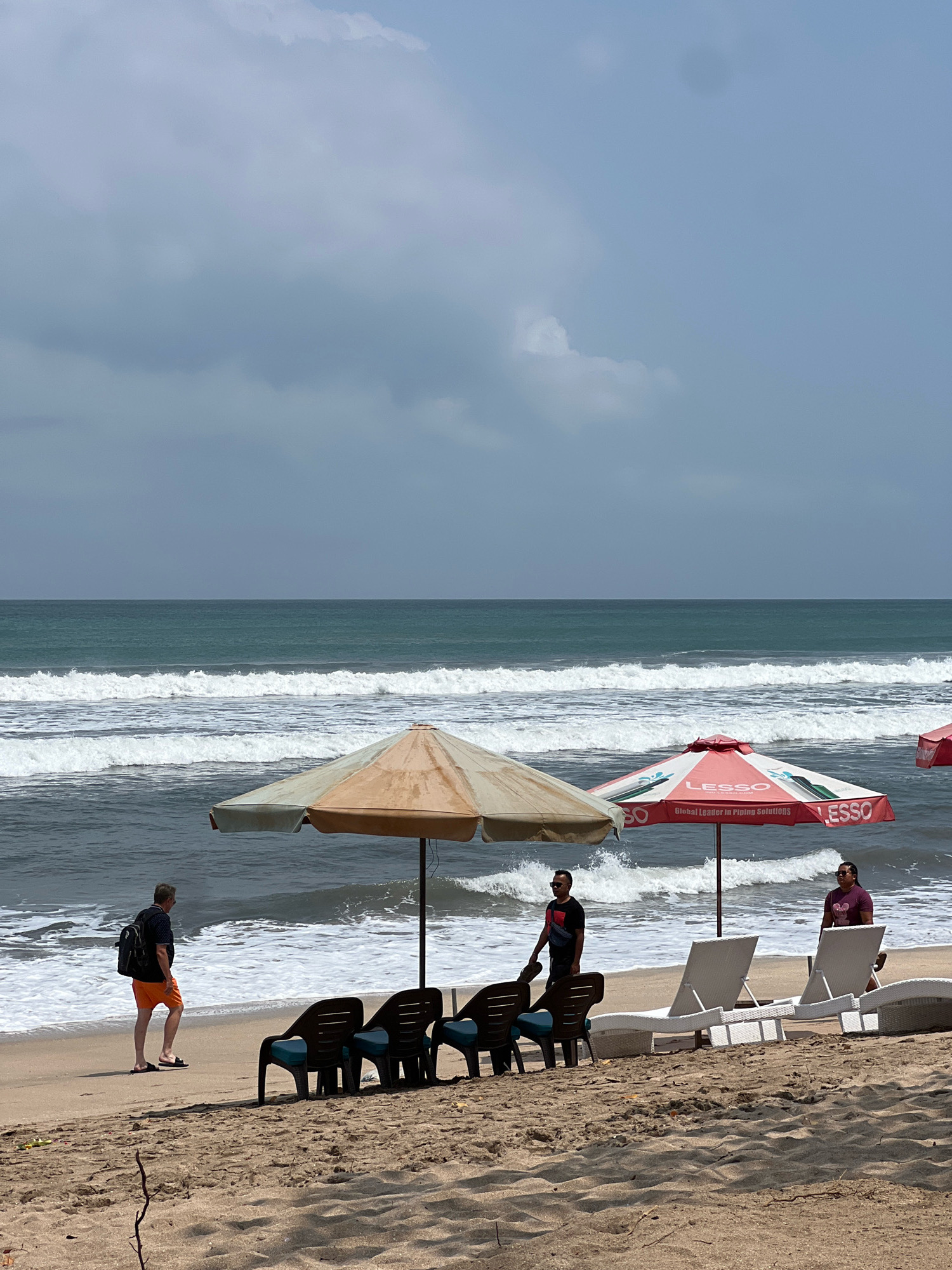 The image depicts a beach scene in Kuta, Indonesia, under a cloudy sky. The ocean is active with moderate waves breaking on the shore. In the foreground, there are several empty beach chairs and two large umbrellas providing shade. One umbrella is red and white, branded with "LESSO Global Leader in Piping Solutions," and the other is a muted beige and green.
Three individuals are present on the beach. On the left, a man wearing orange shorts and a dark patterned shirt walks away from the camera with a black backpack. In the center, a man in a black t-shirt and dark shorts stands near the red umbrella. To the right, a person in a purple t-shirt and dark shorts stands near the white lounge chairs. The sand on the beach is light brown, and there are a few scattered twigs. The overall atmosphere suggests a quiet moment on a popular tourist beach.