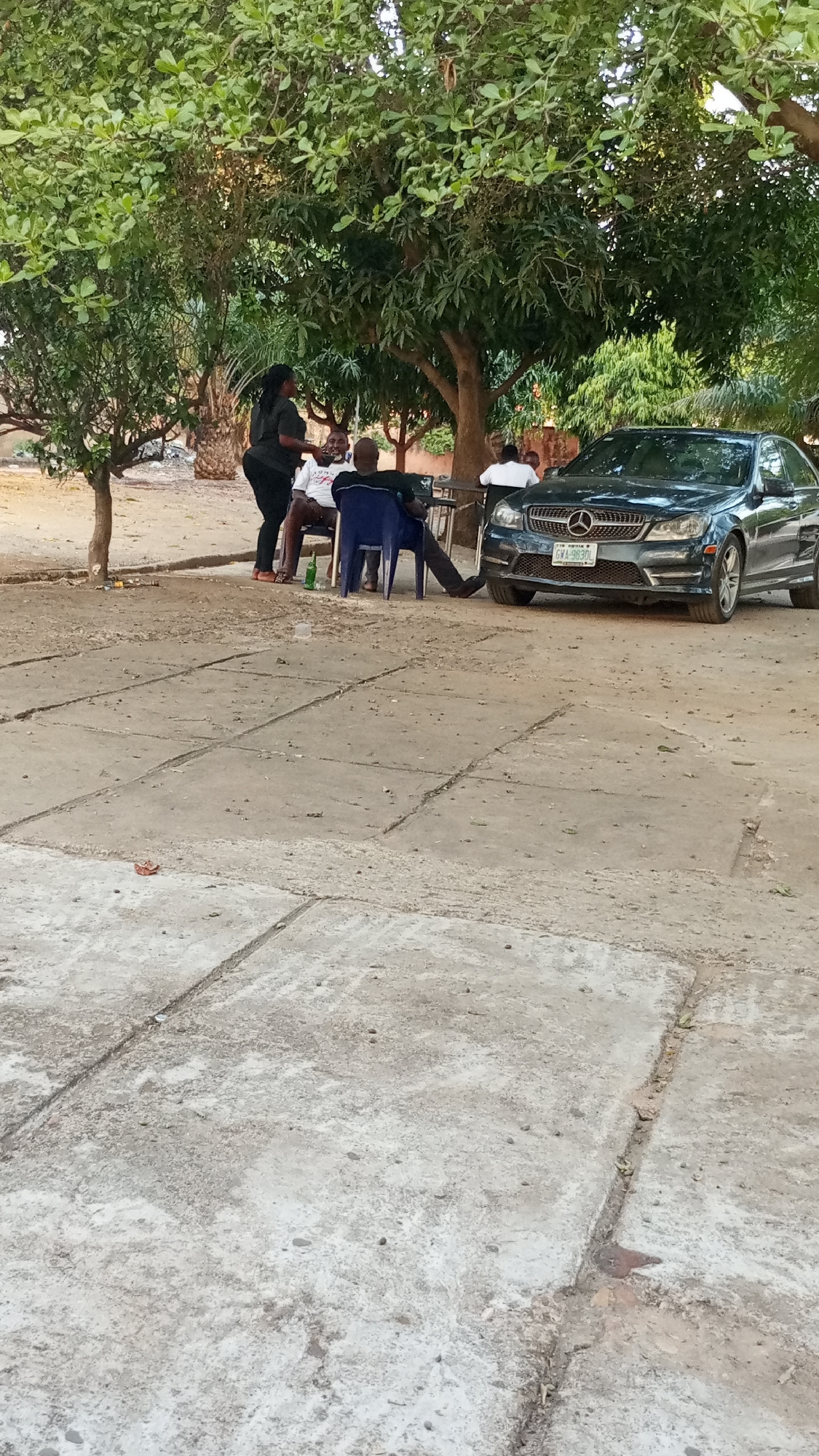 The image shows an outdoor scene in Adankolo, Nigeria. Four people are gathered under trees in a dusty, unpaved area of the city. A woman in black is standing and gesturing toward two men sitting on blue plastic chairs at a small table. A fourth man is seated near the car. A dark-colored Mercedes-Benz with license plate number GWA-983D is parked adjacent to the seating area. There is a green bottle and plastic cup on the ground near the seated men. The ground consists of concrete slabs forming a rough surface. Trees provide shade over the scene.