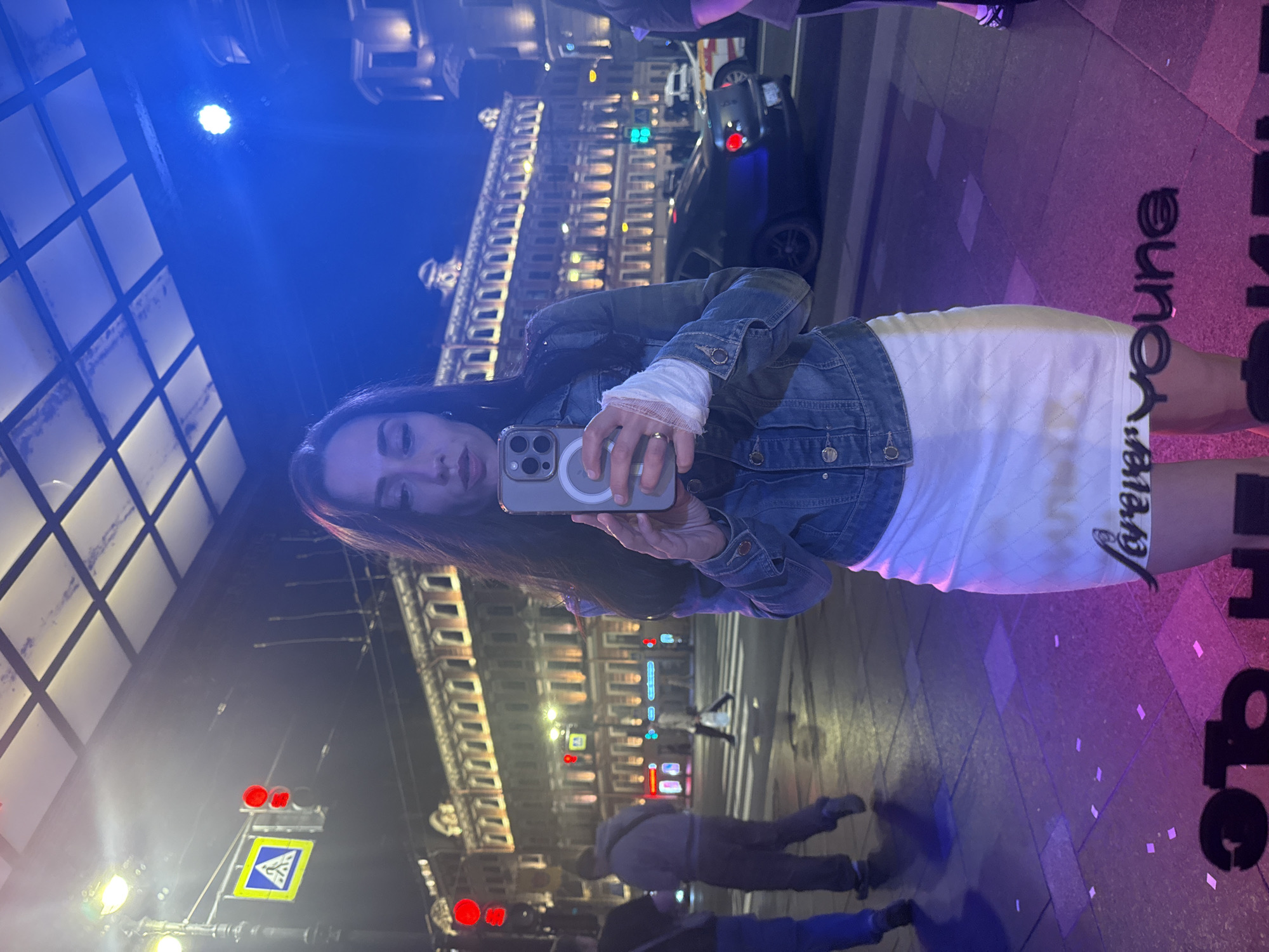 The image is a nighttime selfie captured as a reflection in a window or mirror on a city street in Saint Petersburg, Russia. The main subject is a young woman with long dark hair, holding her smartphone up to take the picture. She is wearing a denim jacket over a white long-sleeved top, which has a white bandage wrapped around her left wrist, and a white textured skirt. A ring is visible on her right hand. Her expression is neutral to slightly serious.
The reflected background reveals a bustling urban scene after dark. Tall, ornate buildings with many illuminated windows, characteristic of Saint Petersburg architecture, line the street. Several cars are visible on the road, along with pedestrians walking on the sidewalk and across a zebra crossing. Traffic lights, including one red and another green, and a blue and yellow pedestrian crossing sign are reflected.
The scene is dramatically lit by various artificial light sources. A bright blue light illuminates the upper left portion, possibly from an interior fixture reflecting outwards. The lower right area is bathed in pink and purple light. On the ground, large, stylized black letters spell out "Forever Young," although the last part is cut off by the edge of the image. The overall setting suggests a lively city evening.