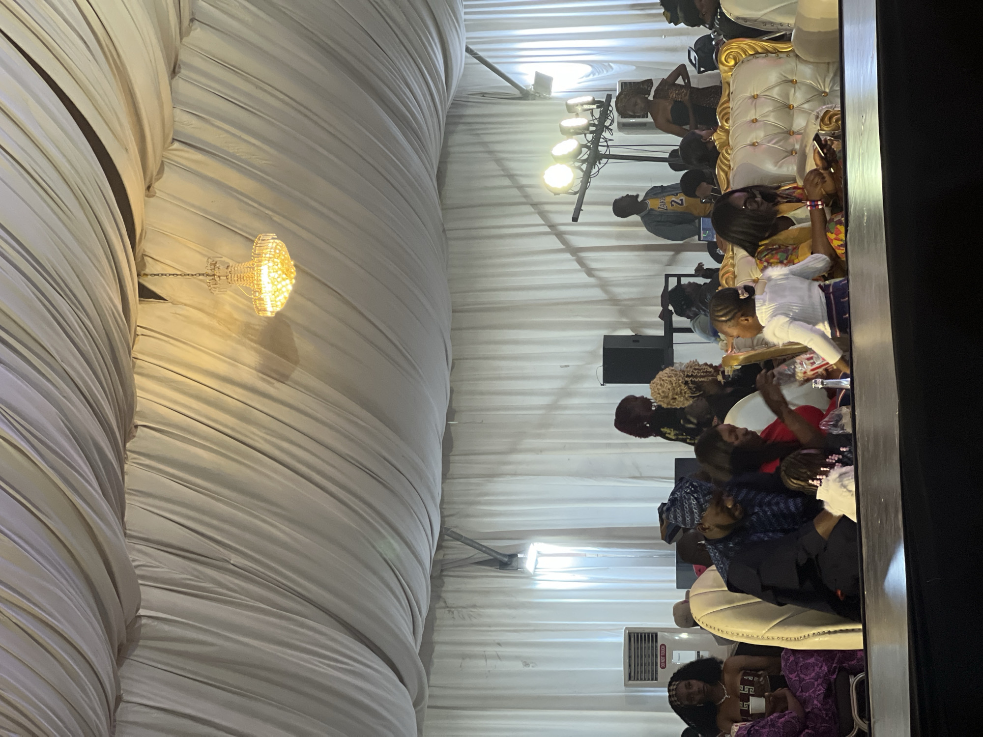 The image depicts an indoor event in Barakin Lerawi, Nigeria, likely a celebration or gathering, held under a draped white tent ceiling. A decorative chandelier hangs from the ceiling. In the foreground, several individuals are seated around tables, some engaged in conversation, others looking at their phones. A woman in a yellow and multi-colored patterned outfit is seated and looking at her phone, with a child in a white top and striped skirt to her left. To her right, a woman in a red top is also seated. In the middle ground, a man in a dark blue patterned cap and matching attire is seated and looking down, possibly at a child near him. Other guests are visible in the background, some standing. Among the standing figures, a man wearing a yellow "Lakers" jersey is noticeable. The environment appears to be a banquet hall or event space, with white drapes covering the walls and ceiling, and various lighting fixtures, including spotlights on a stand. An air conditioning unit is visible on the left side of the frame. The overall atmosphere suggests a social event with an audience.