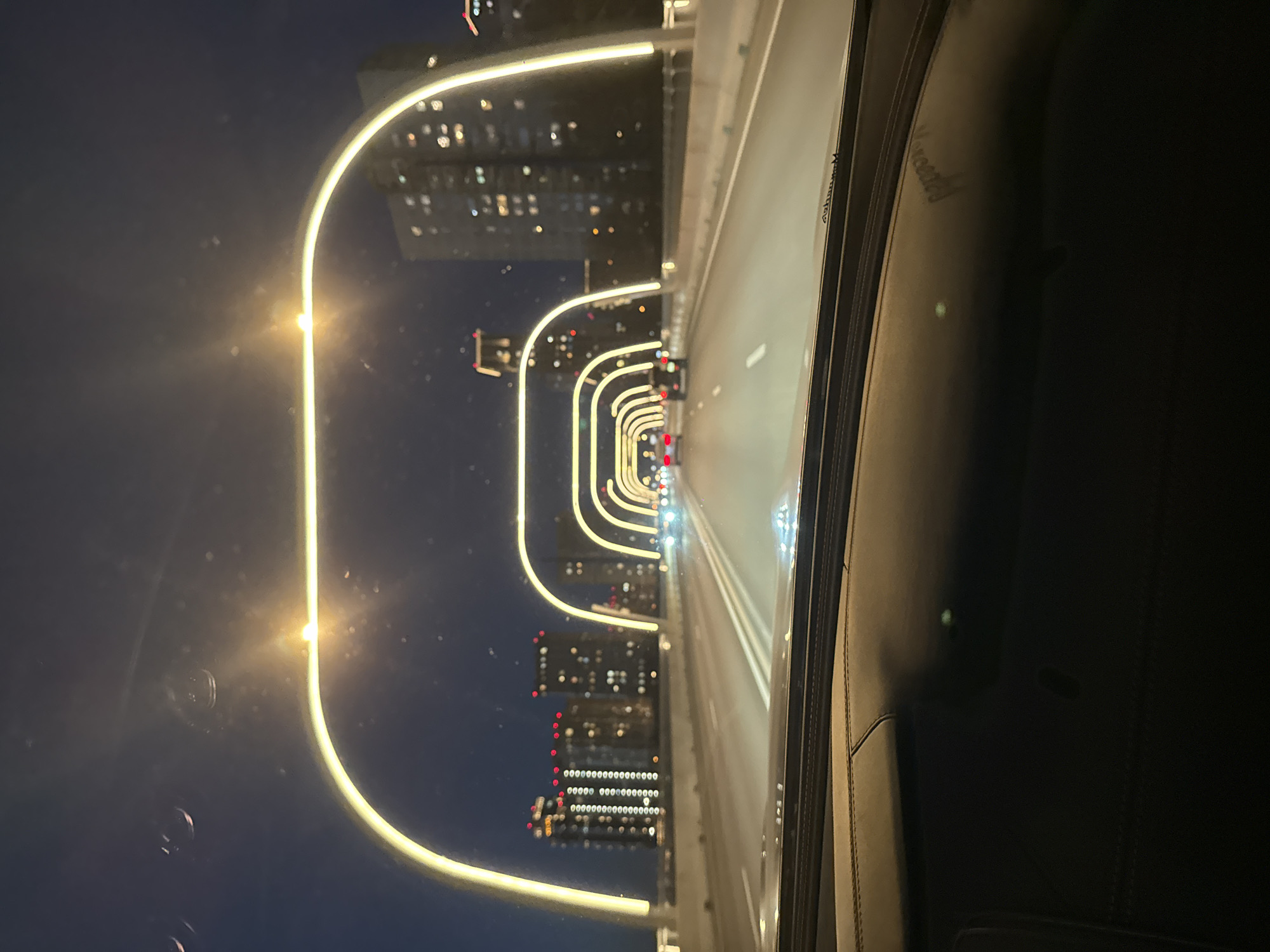 The image displays a nocturnal scene captured from inside a car, looking out onto a modern highway or bridge in what is likely Khimki, Russia. The photograph is oriented sideways (rotated 90 degrees counter-clockwise), with the road appearing vertically.
The central subject is a multi-lane road dramatically illuminated by a sequence of bright, arched streetlights. These lights line both sides of the thoroughfare, creating a distinctive, tunnel-like effect that extends into the vanishing point. In the background, against the dark night sky, numerous illuminated windows of distant tall buildings are visible, indicating an urban or suburban environment.
The foreground on the right side shows the dark interior of the vehicle, including a portion of the dashboard or door panel with visible stitching. Faint reflections on the car window reveal stylized text. The word "Maserati" is clearly discernible, with another, less distinct word above it, possibly "Monterosso," suggesting the make of the vehicle.
The activity depicted is driving along this uniquely lit highway at night. The weather appears clear, with no visible precipitation or fog.