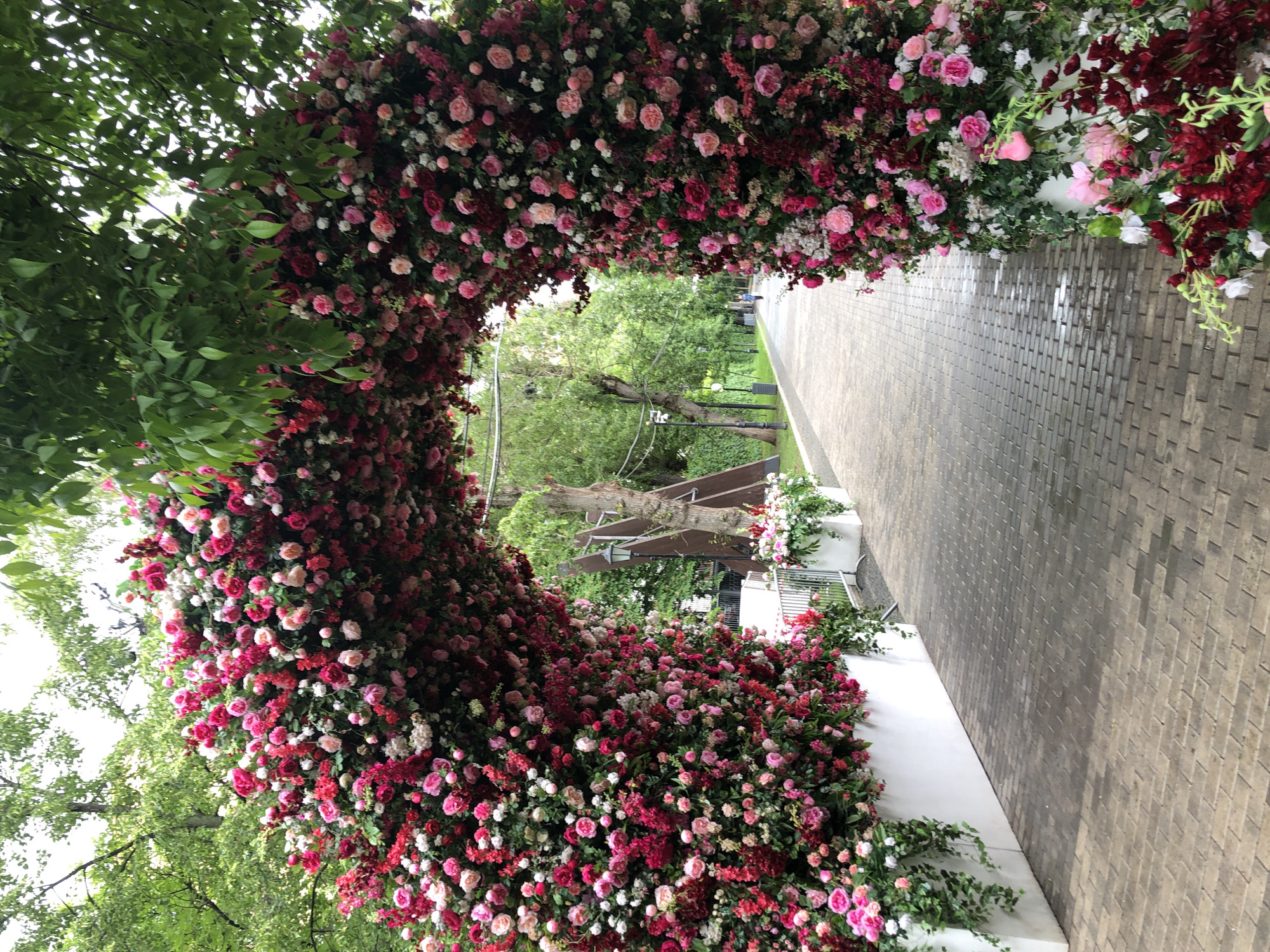 The image shows a walkway in what appears to be a park or garden, possibly in Moscow, Russia. The walkway is lined with brick or stone paving. A large archway covered in red, pink, and white roses is constructed over the walkway. Green foliage is visible amongst and beside the roses, adding to the garden setting. In the distance, there are trees and a lamp post, suggesting a public space. A wooden structure with floral decorations is partially visible on the right side of the image. No people are visible in the immediate scene.